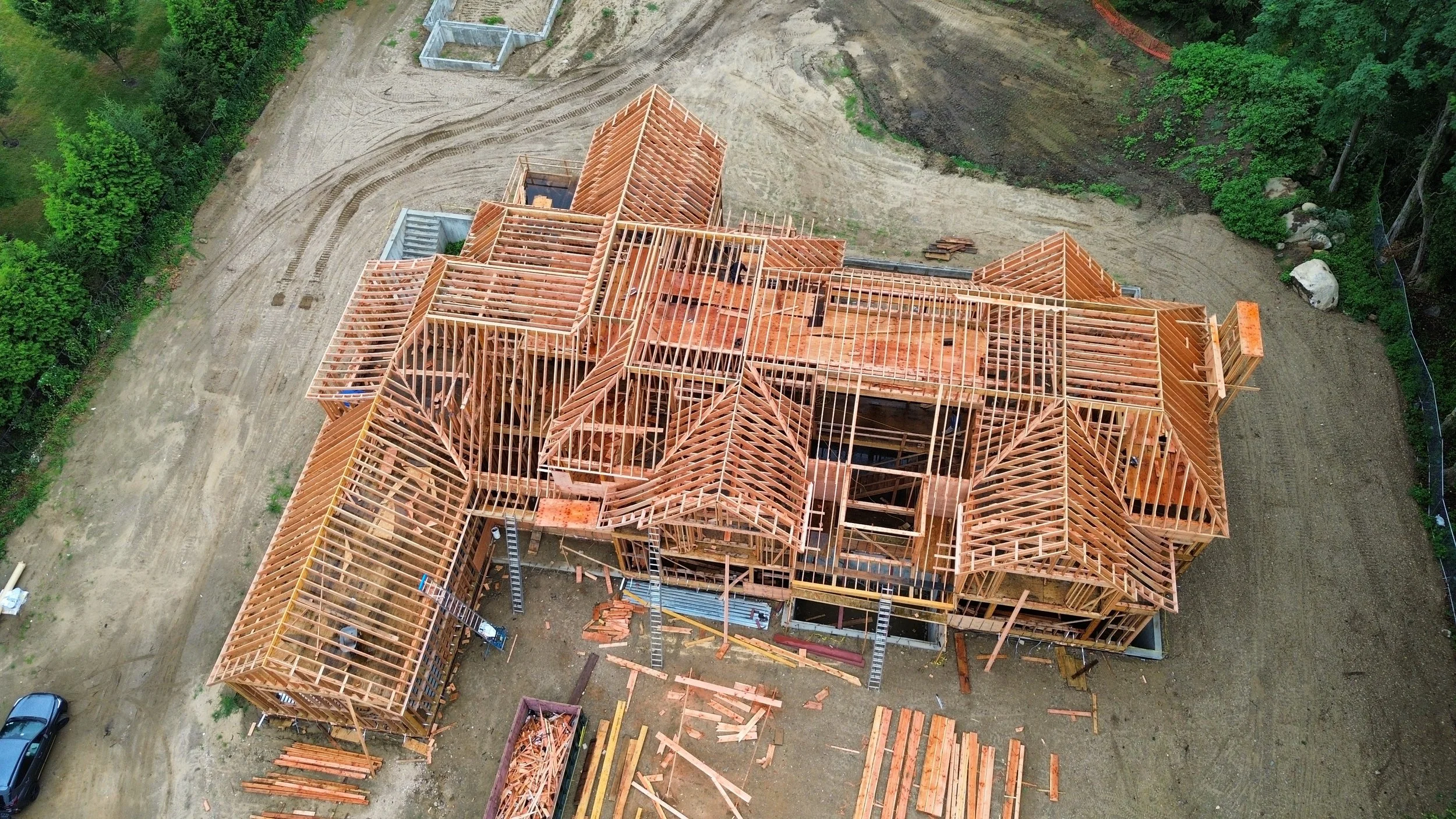 An aerial view of a house under construction with a wooden frame and roof structure, surrounded by dirt and greenery.