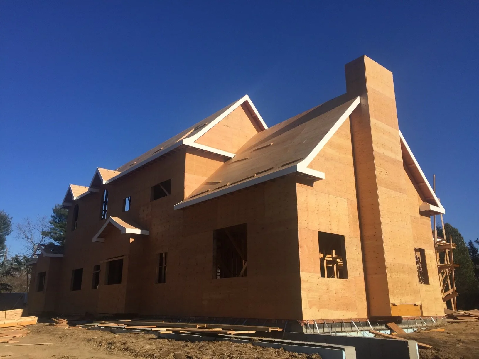 A house under construction with wooden framing and roofing, set against a clear blue sky.