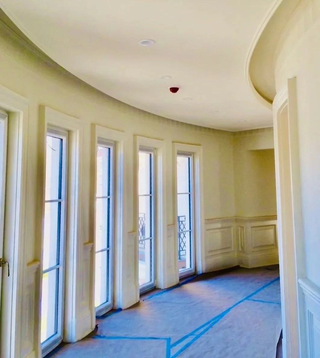 Empty room with large curved windows, cream-colored walls, and hardwood floor under construction.