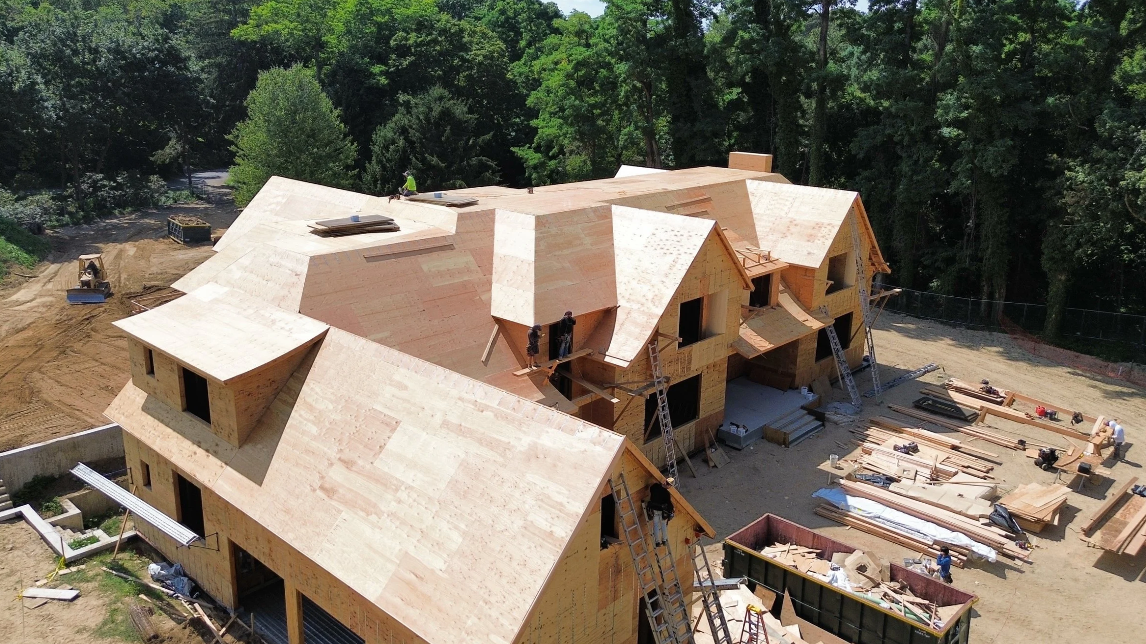 Aerial view of a large house under construction with wooden framing, workers on site, and surrounded by trees.