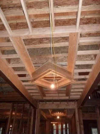 A shot of an unfinished ceiling in a building under construction with exposed wooden beams and framing, featuring a modern geometric light fixture hanging in the center.