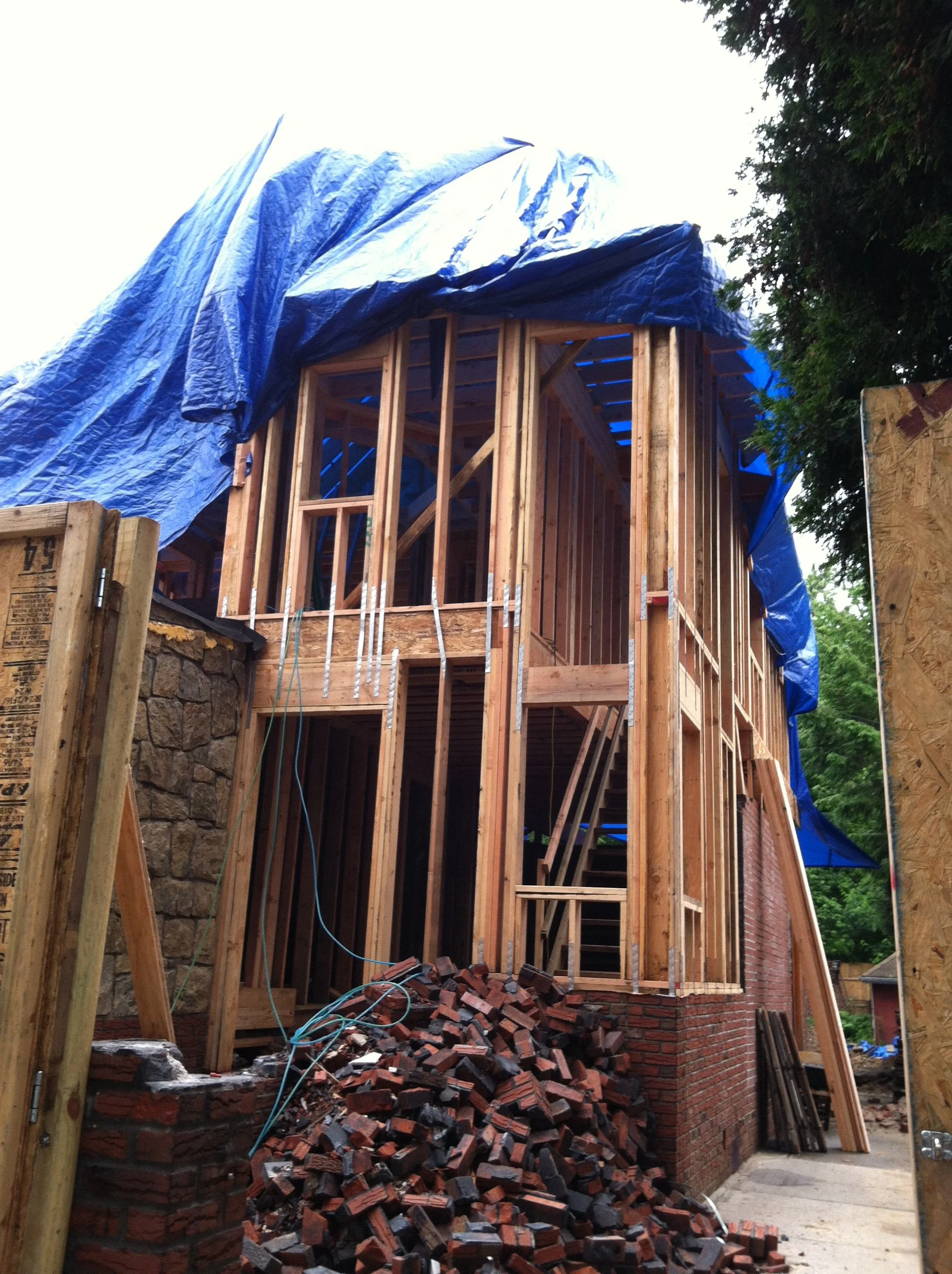 Construction site showing a partly built multi-story residential building with wooden framing, scaffolding, and a pile of red bricks in front.