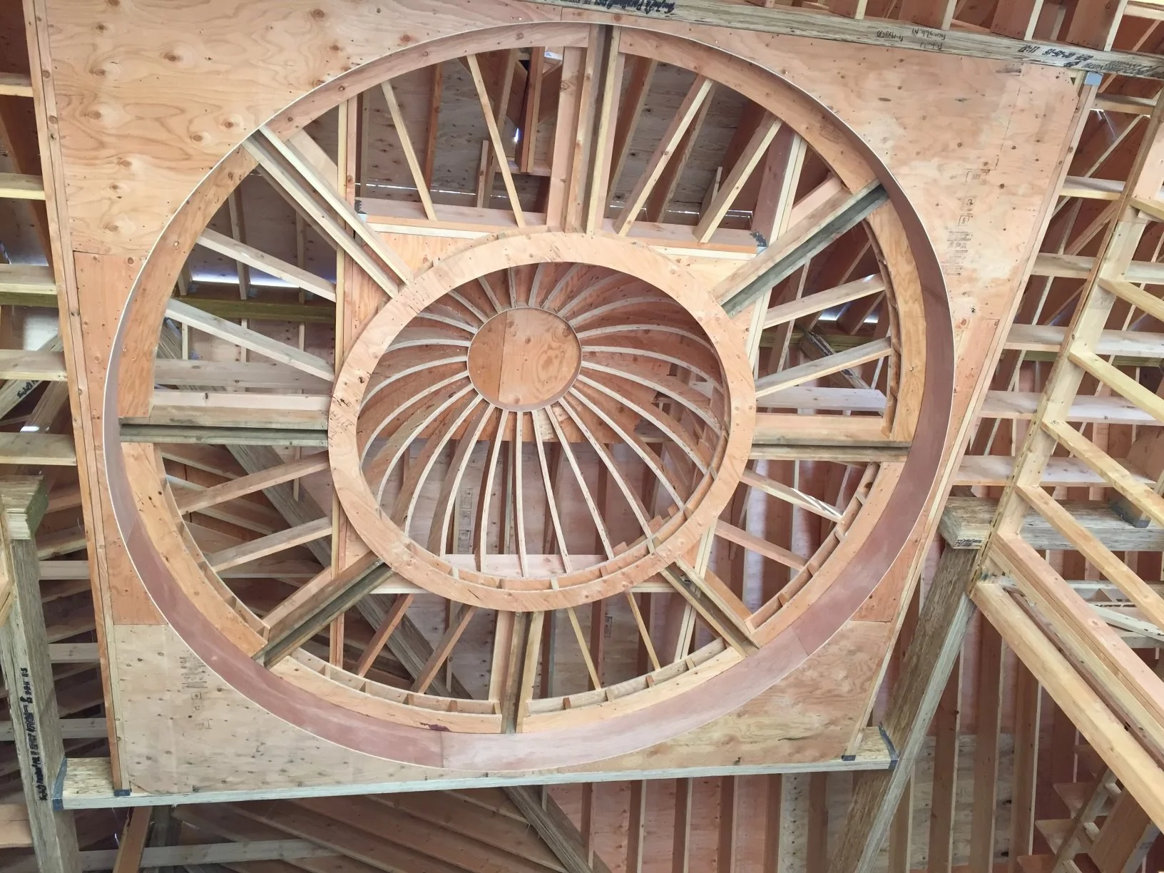Wooden spiral staircase framework under construction, showing concentric circles and radial supports, within a building under construction.