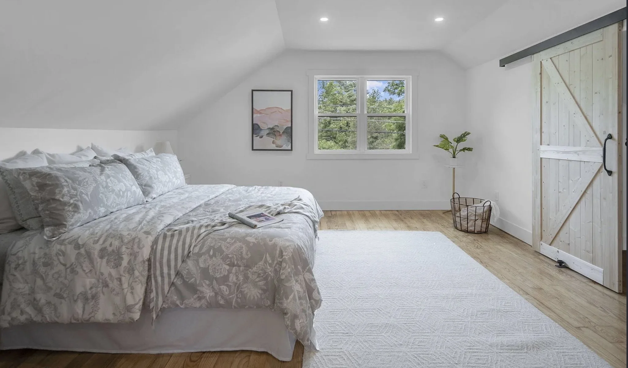 A bedroom with a bed on the left, a window showing green trees outside in the center, and a sliding barn door on the right. The room has white walls, a wooden floor, a white rug, a small plant, and a book on the bed.
