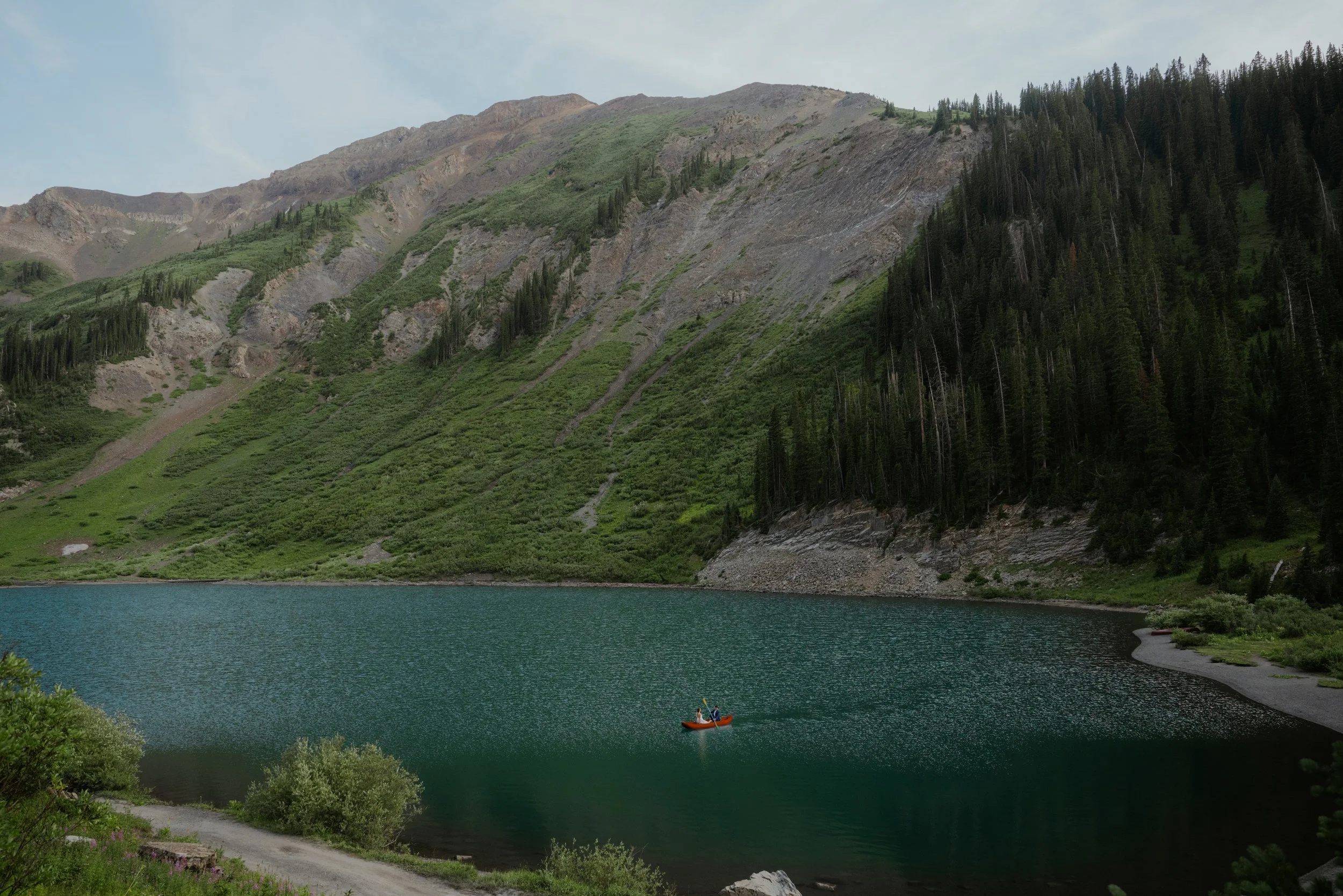 A lake surrounded by green hills and mountains with a small boat on the water and a trail along the shoreline.