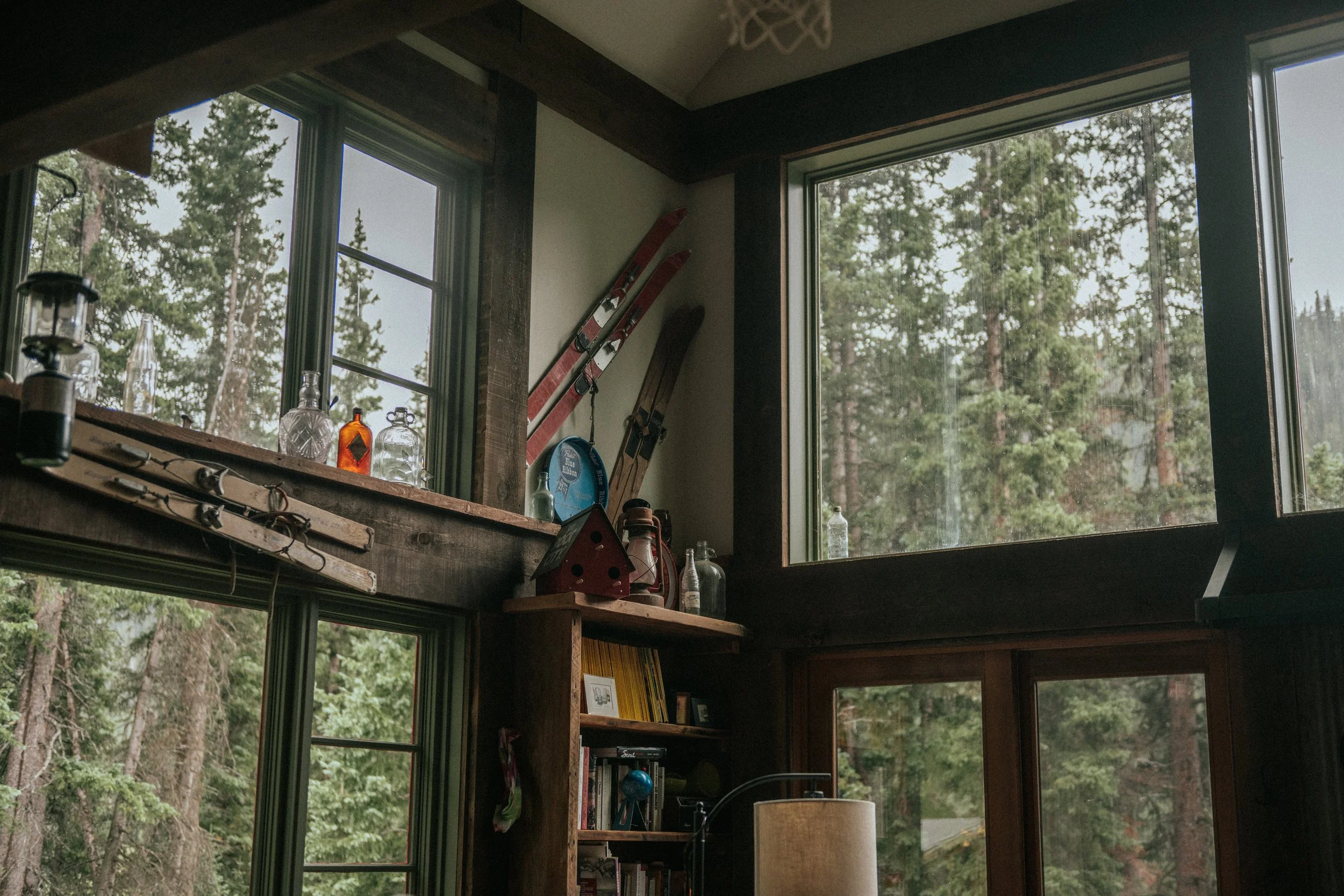 Interior of a rustic cabin with large windows showing a forest outside. Inside, there are vintage bottles on a wooden shelf, skis leaning against the wall, and a bookshelf with books and decor items.