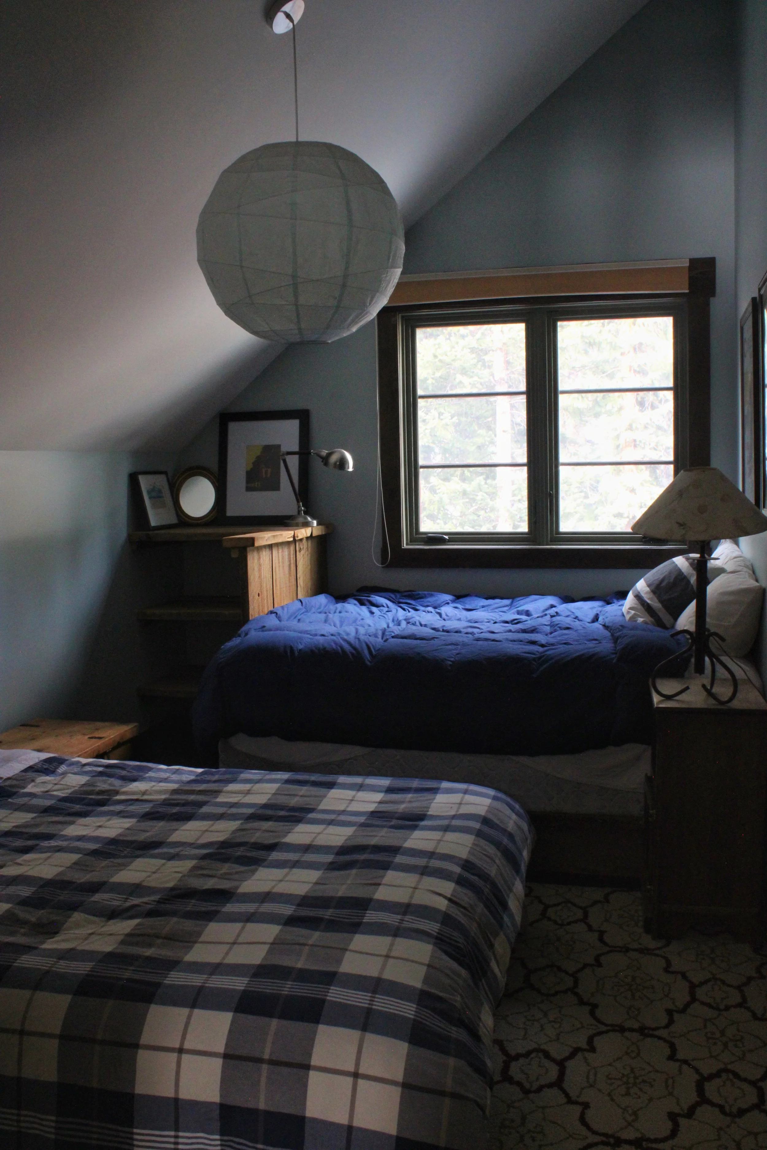 A bedroom with two beds, one with a plaid blanket and the other with a solid blue blanket. There is a window with a view of trees outside. The room has a sloped ceiling, a wooden shelf with picture frames, a lamp, and a patterned rug on the floor. A paper lantern hangs from the ceiling.