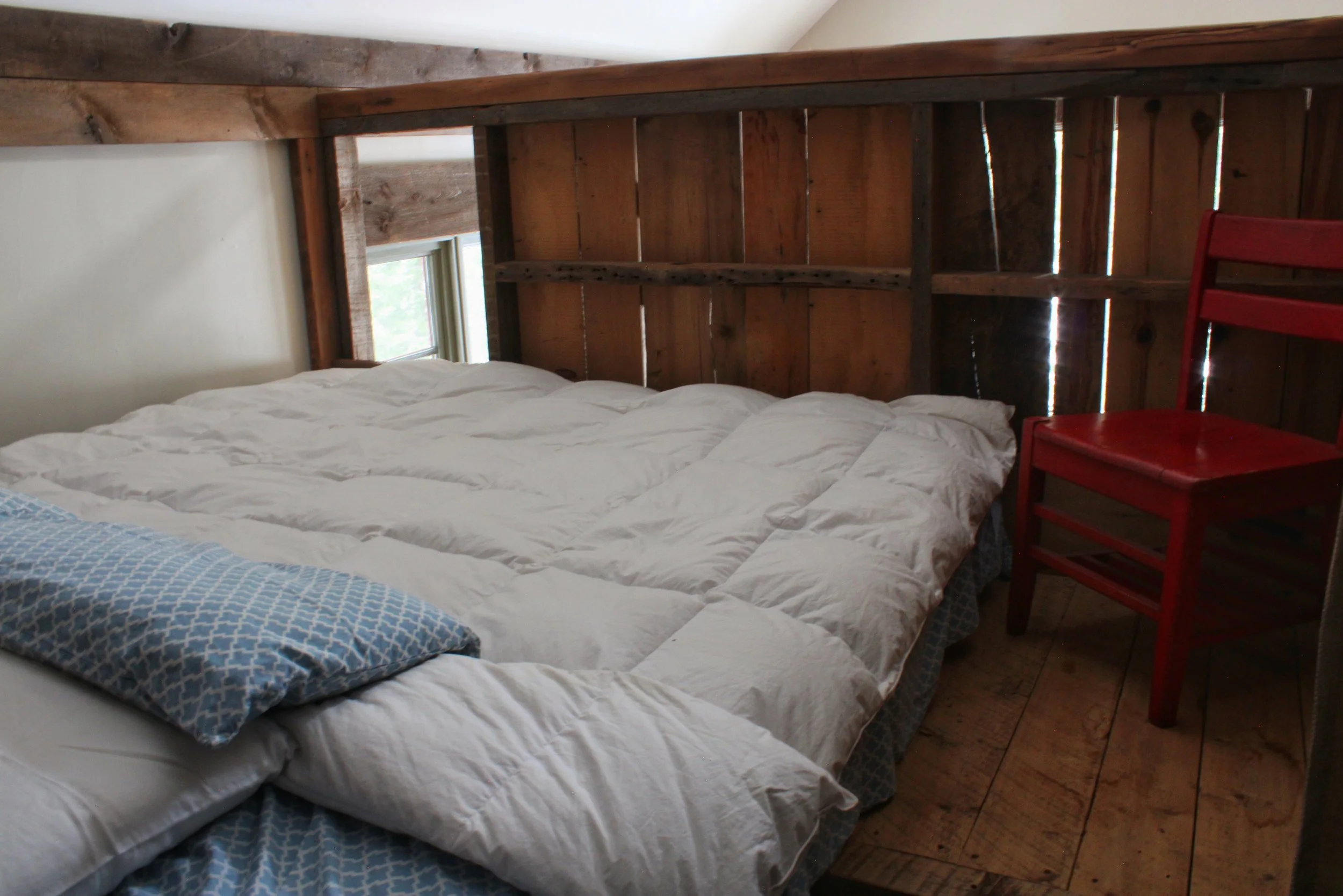 A cozy bedroom with a white bed and a blue patterned pillow. Wooden headboard and side table, with a small window providing natural light. Wood-paneled walls and flooring add warmth to the space.
