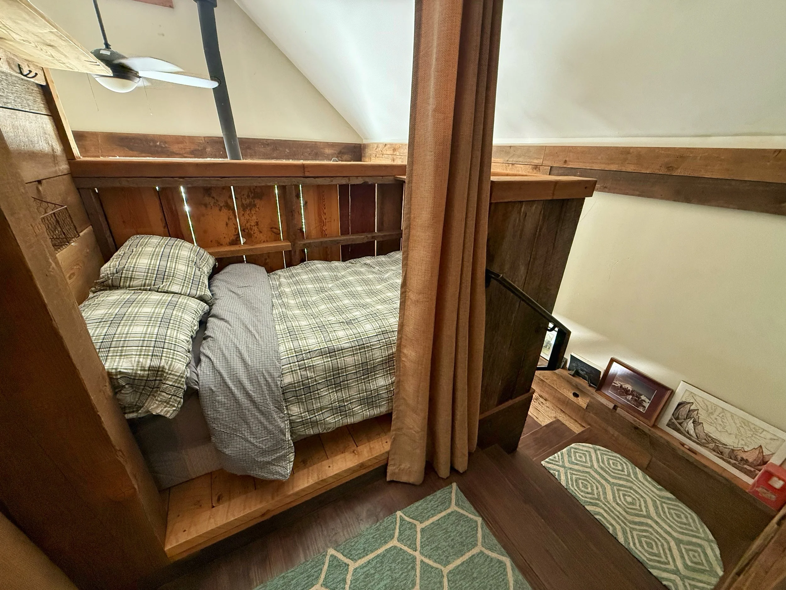 A cozy bedroom with wooden furniture, including a bed with plaid bedding, a framed picture on the floor, and a hanging curtain partially obscuring the room. The walls are cream-colored, and the floor is dark wood.