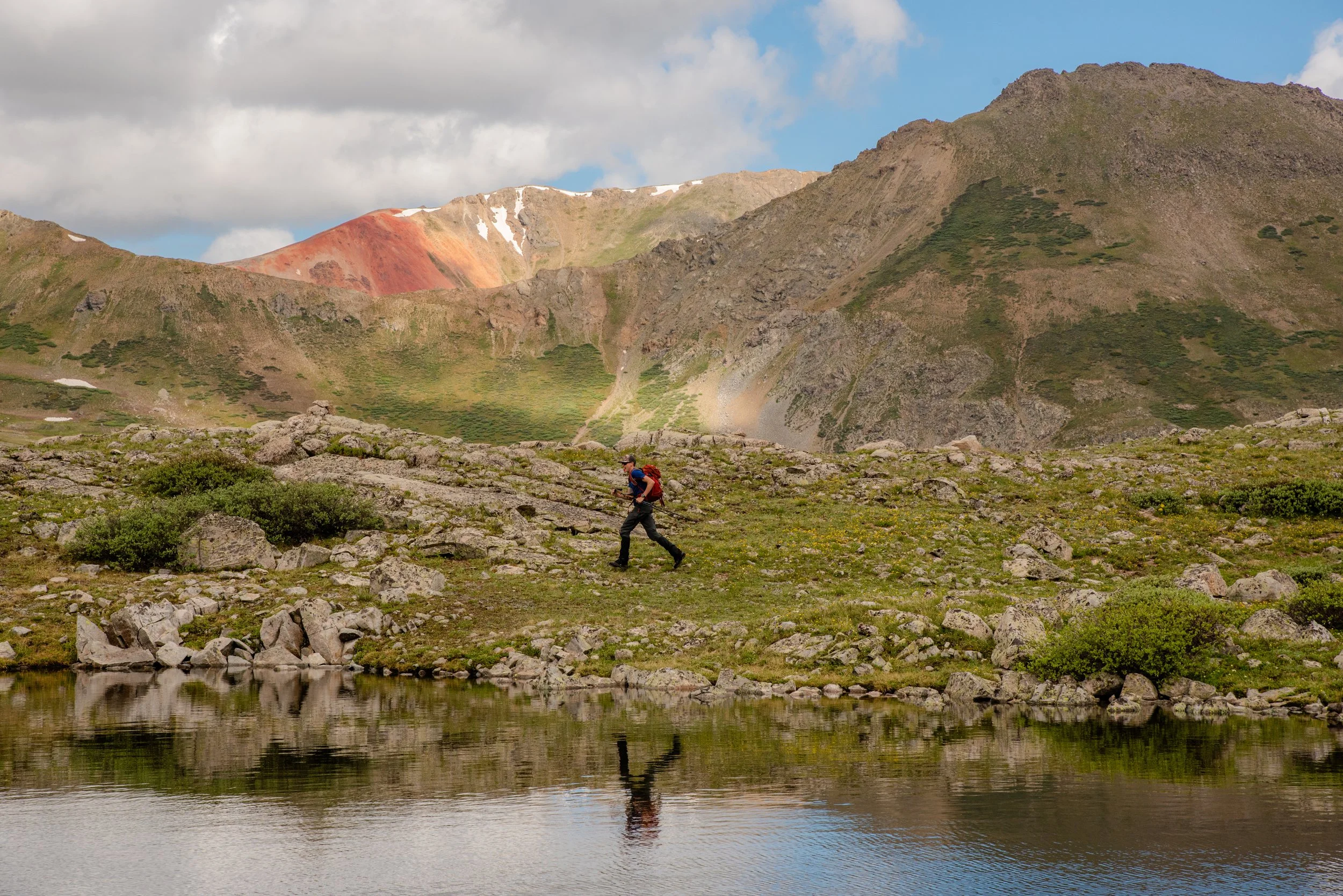 Hiker wearing a blue shirt and a red backpack walking on rocky terrain near a body of water with mountains in the background.