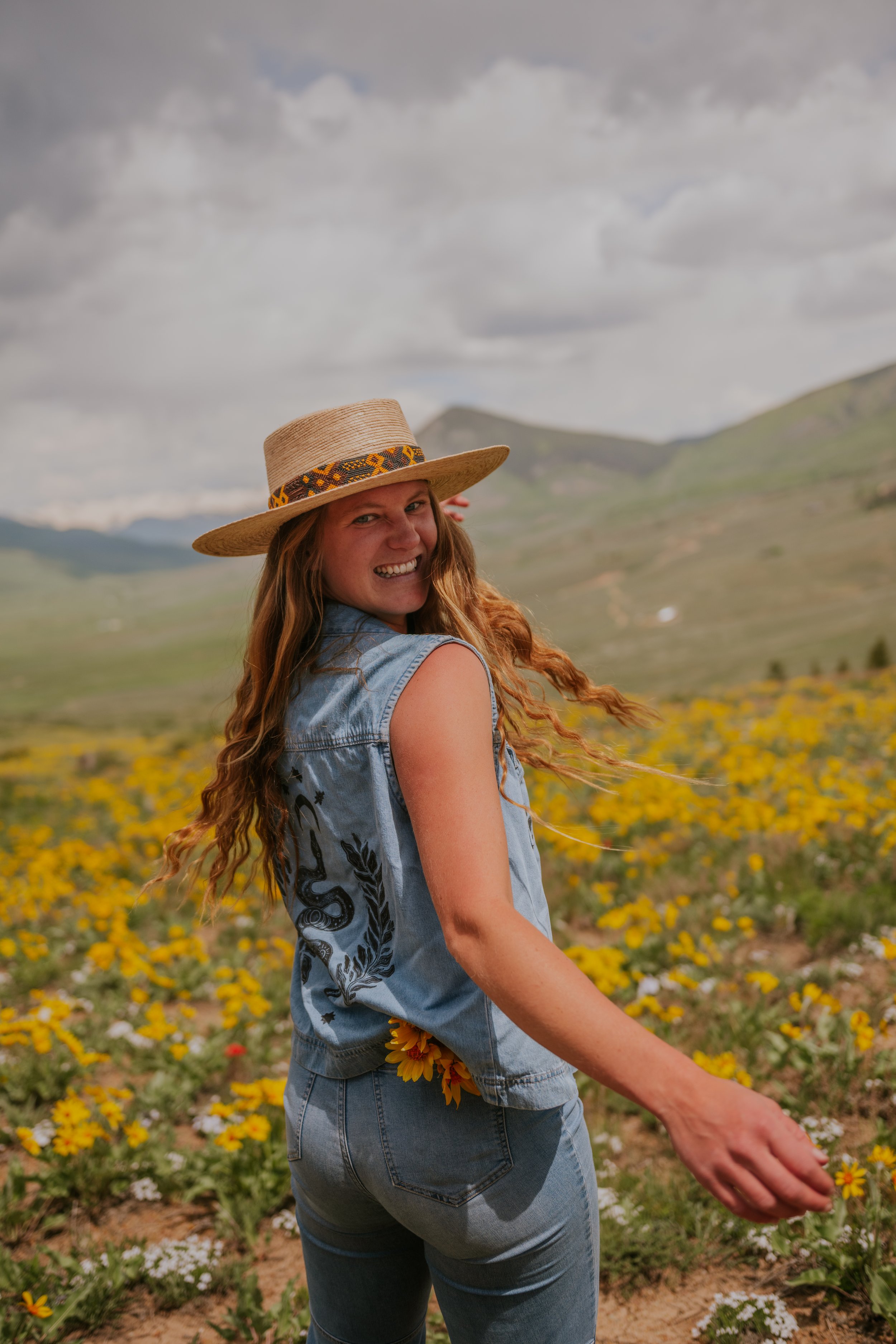 A woman with long, curly red hair wearing a wide-brimmed straw hat and a sleeveless denim vest, standing in a field of yellow and white flowers with mountains in the background, under a cloudy sky, smiling at the camera.