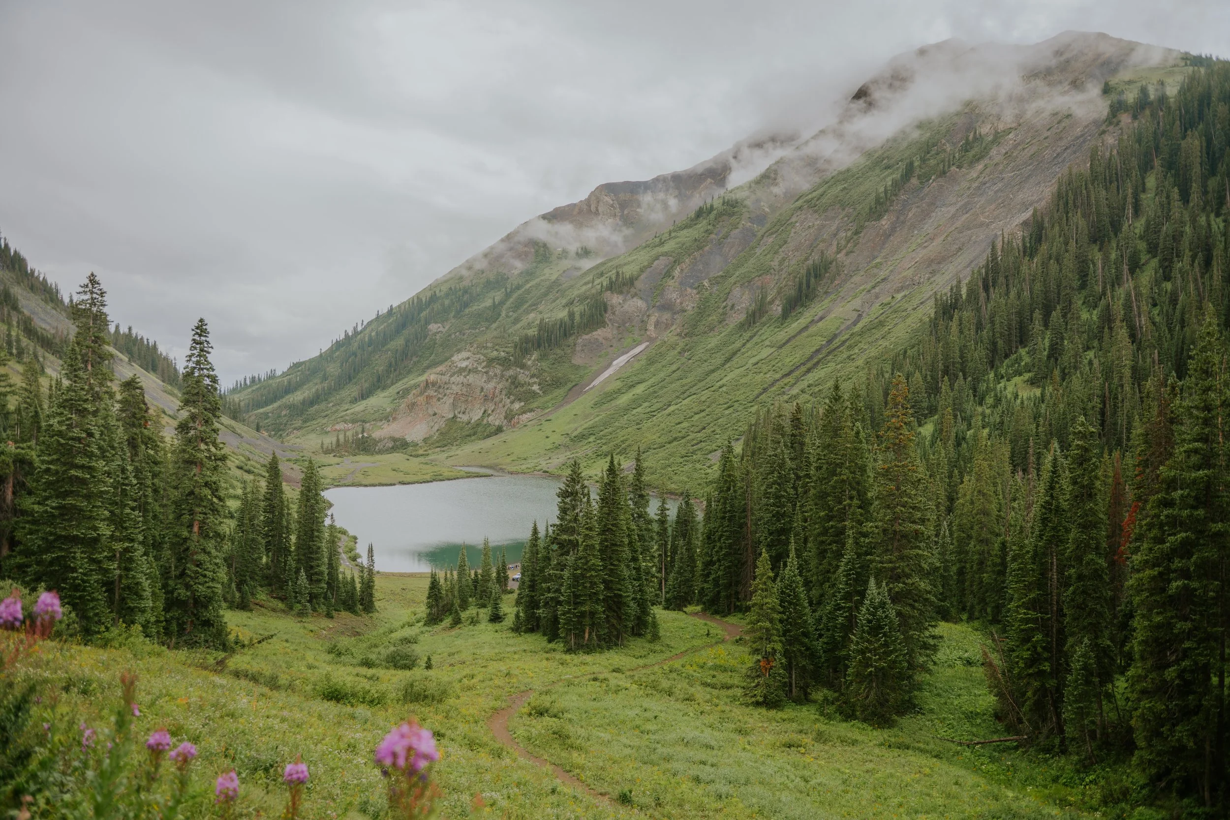 A mountainous landscape with green forests, a small lake, and misty clouds covering the mountain peaks.