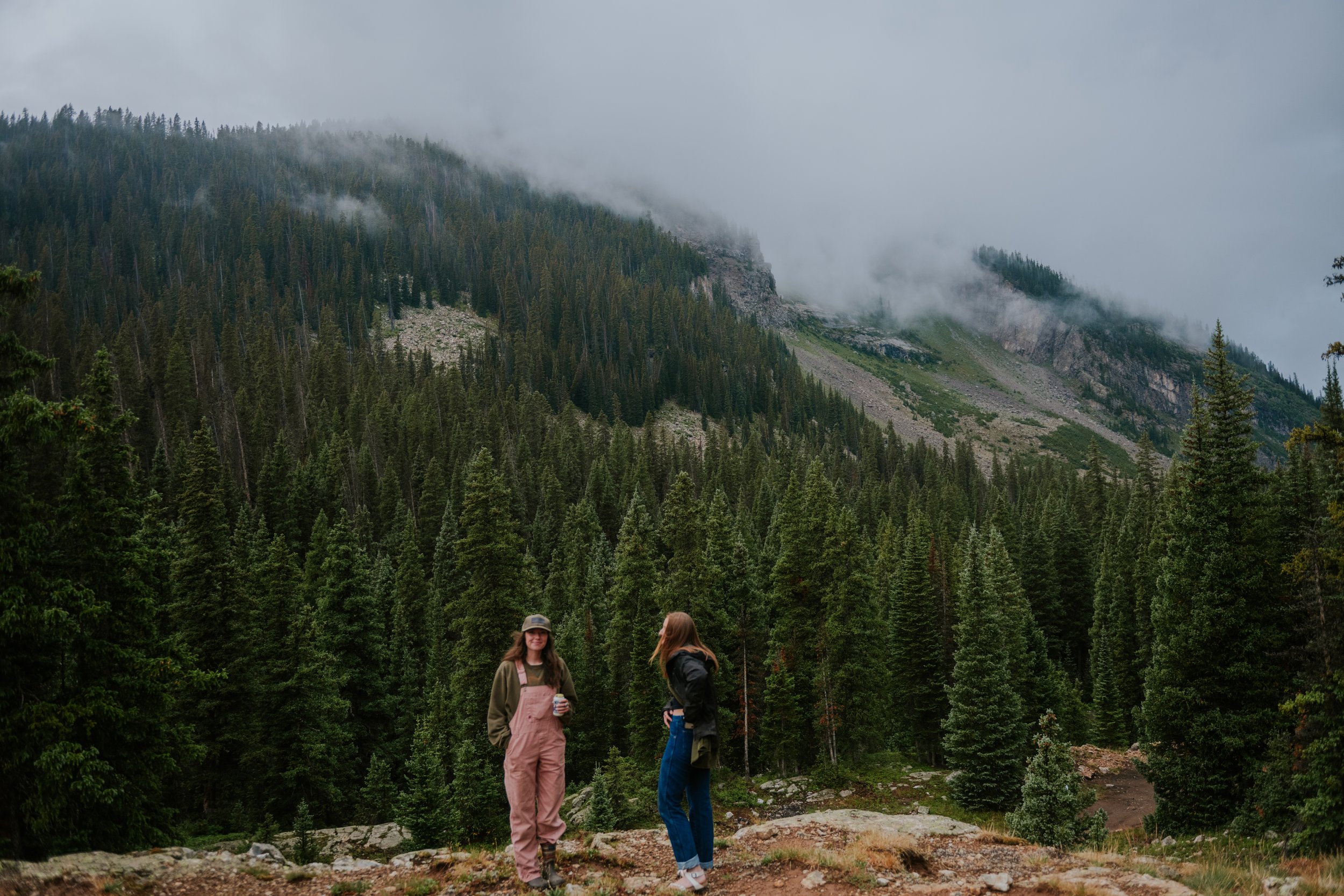 Two women stand in a forested mountain area with dense evergreen trees, rocky terrain, and mist-covered mountain slopes in the background.