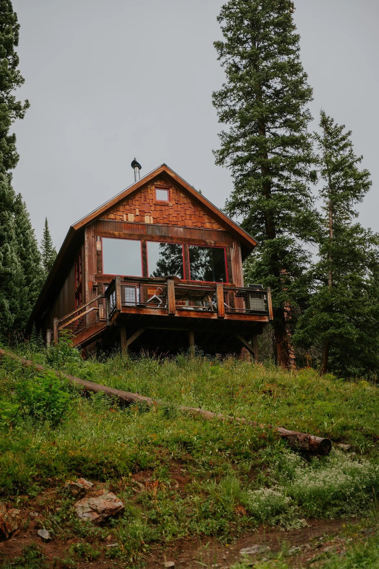 A wooden house with a balcony surrounded by tall trees and a green grassy hillside.