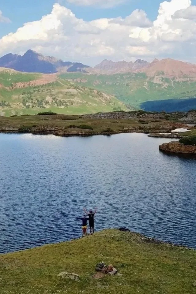 Two people standing on a grassy bank beside a lake, with mountains and a cloudy sky in the background.