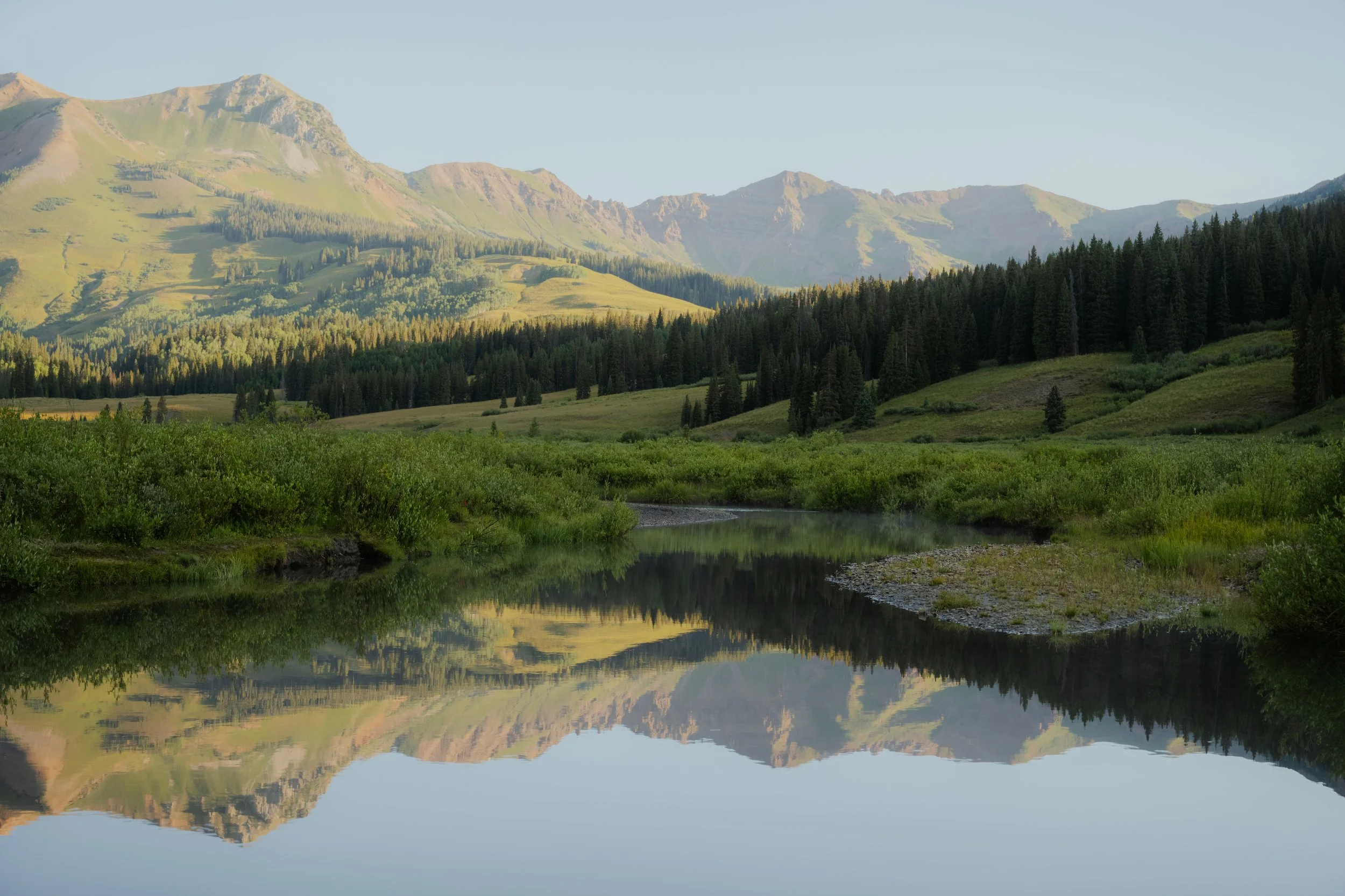 A serene mountain landscape with a calm river reflecting the green hills, dense forest, and mountain peaks under a clear sky.