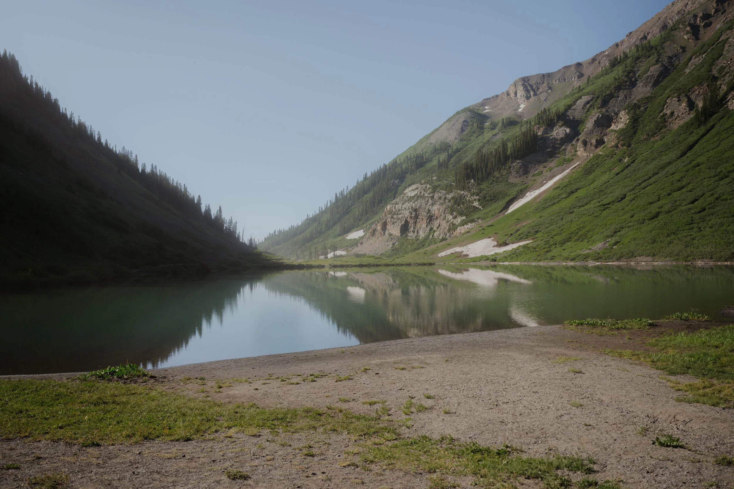 A serene mountain landscape with a calm lake reflecting green, rocky mountain slopes on both sides under a blue sky.