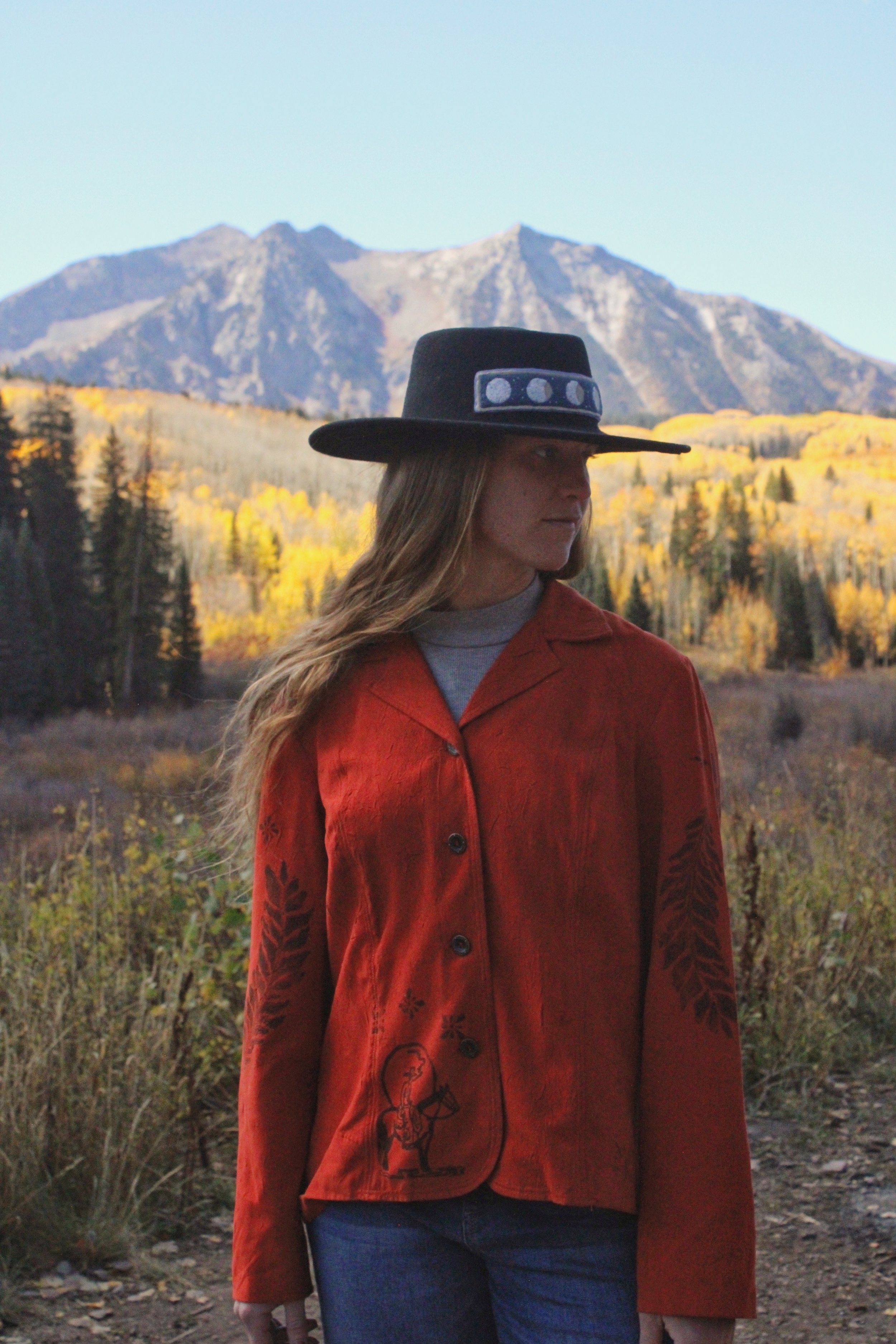 A woman wearing a black hat with a band of circular designs, a red jacket with black embroidery, and blue jeans, standing outdoors with a mountain in the background. The mountain is surrounded by trees with yellow and green leaves, indicating autumn.