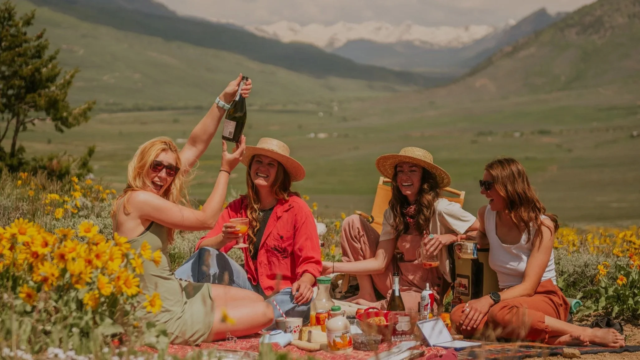 A group of four women having a picnic outdoors in a field of yellow flowers with mountains in the background. One woman is holding champagne and others are smiling and enjoying their drinks.
