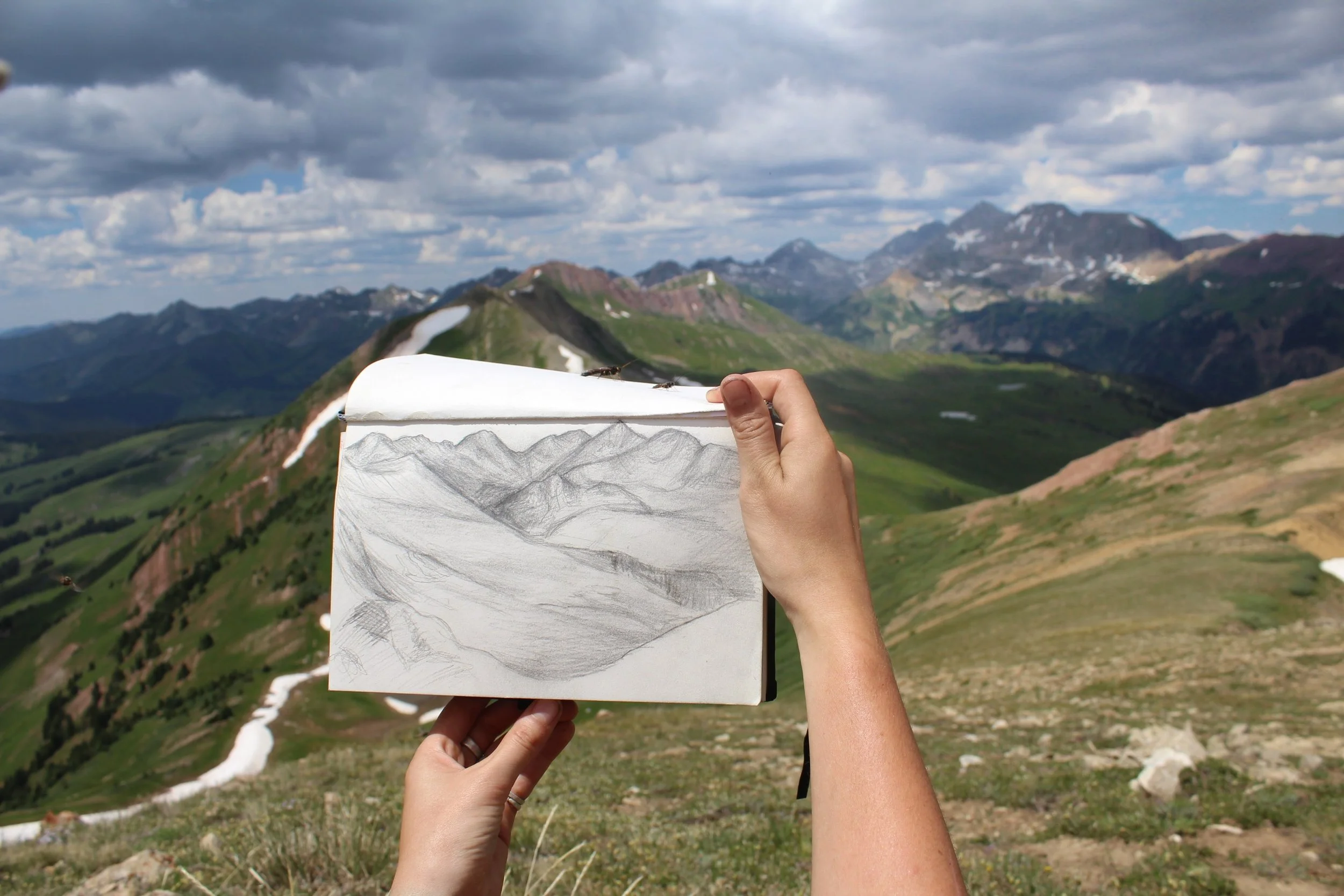 A person holding a sketchbook with a pencil drawing of mountains, with a mountain landscape in the background featuring green mountains, patches of snow, and a cloudy sky.