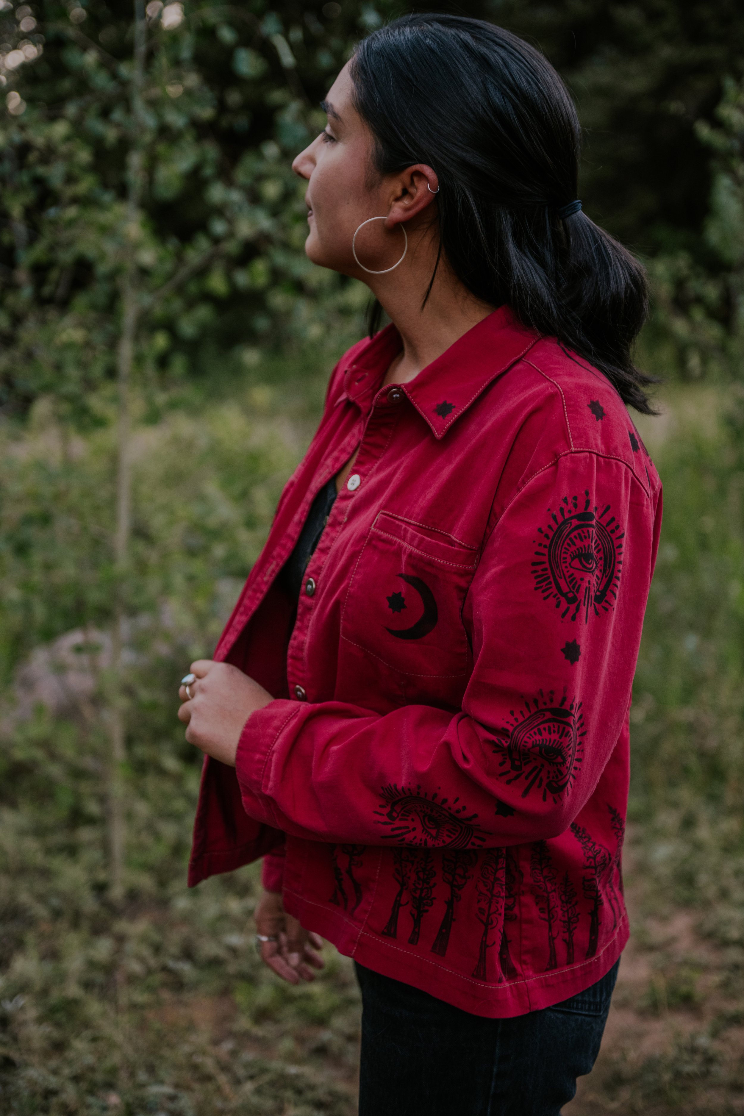 Profile of a woman with dark hair tied back, wearing a red jacket with black printed designs, standing outdoors with green foliage in the background.