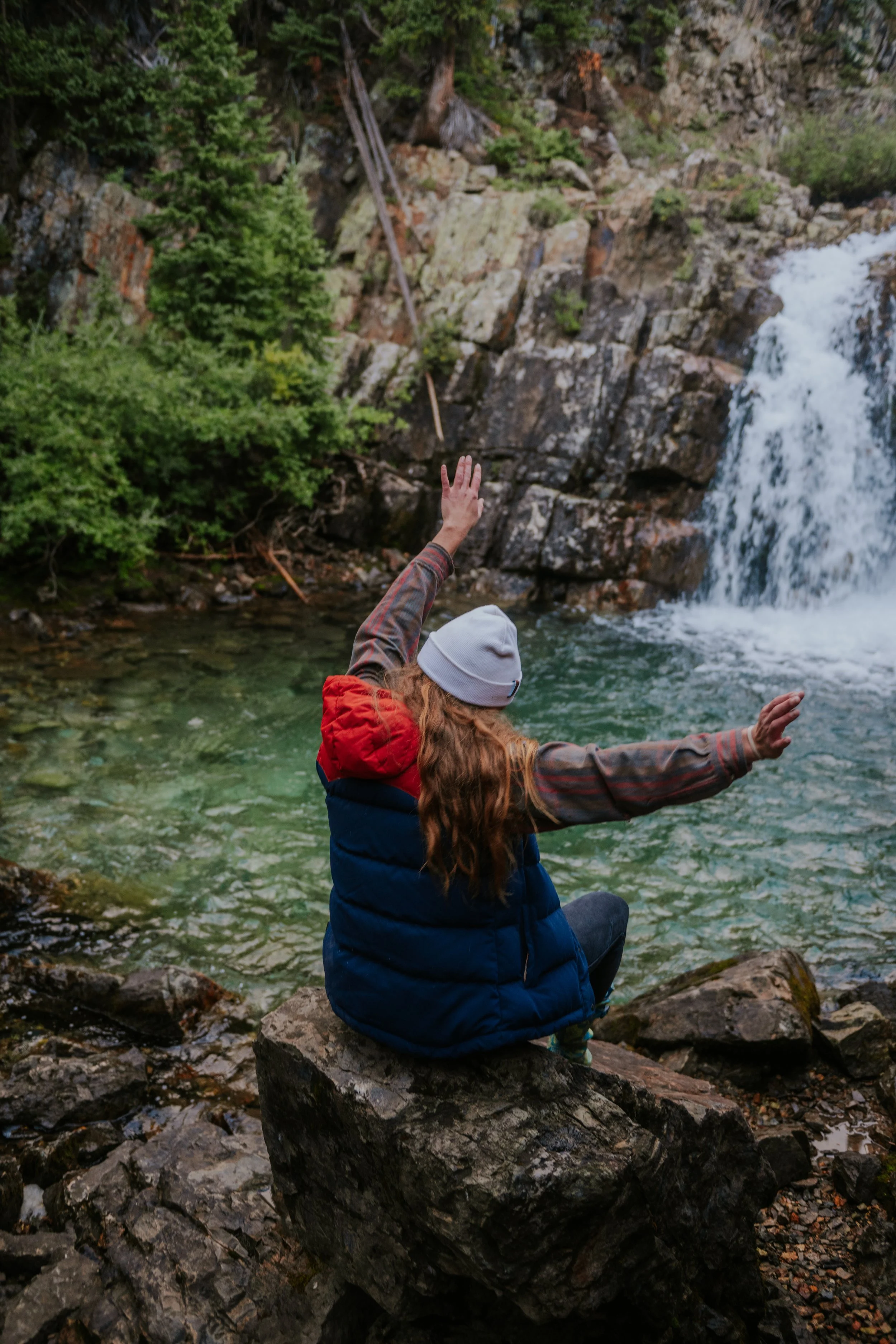 A person sitting on a rock by a river, wearing a grey beanie, a red and blue vest, and a plaid long-sleeve shirt, with one arm raised to throw a rock into the water, near a waterfall surrounded by trees.
