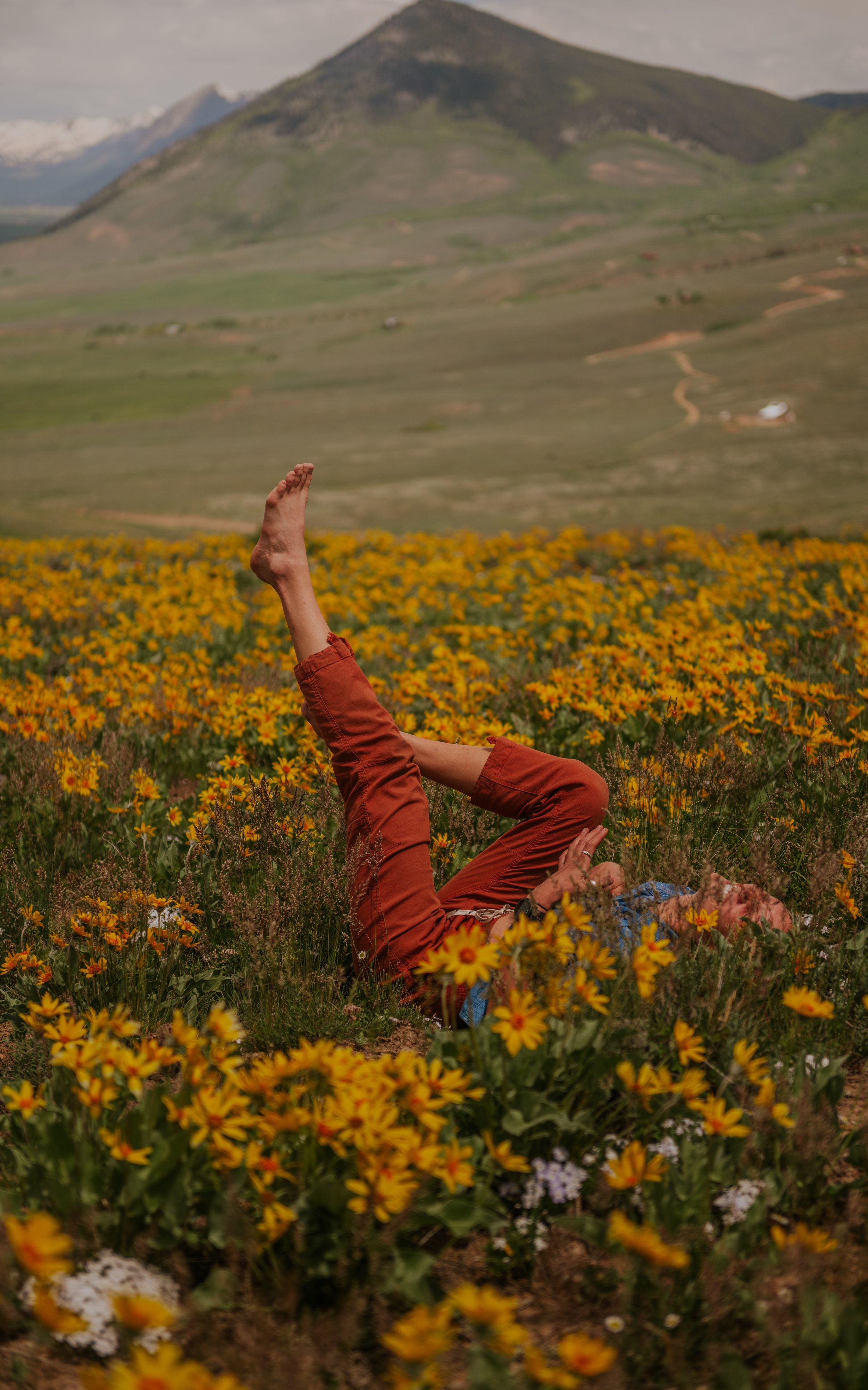 A person lying on the ground in a field of yellow flowers, doing a yoga pose with one leg raised and knees bent, wearing orange pants, with a mountain in the background.