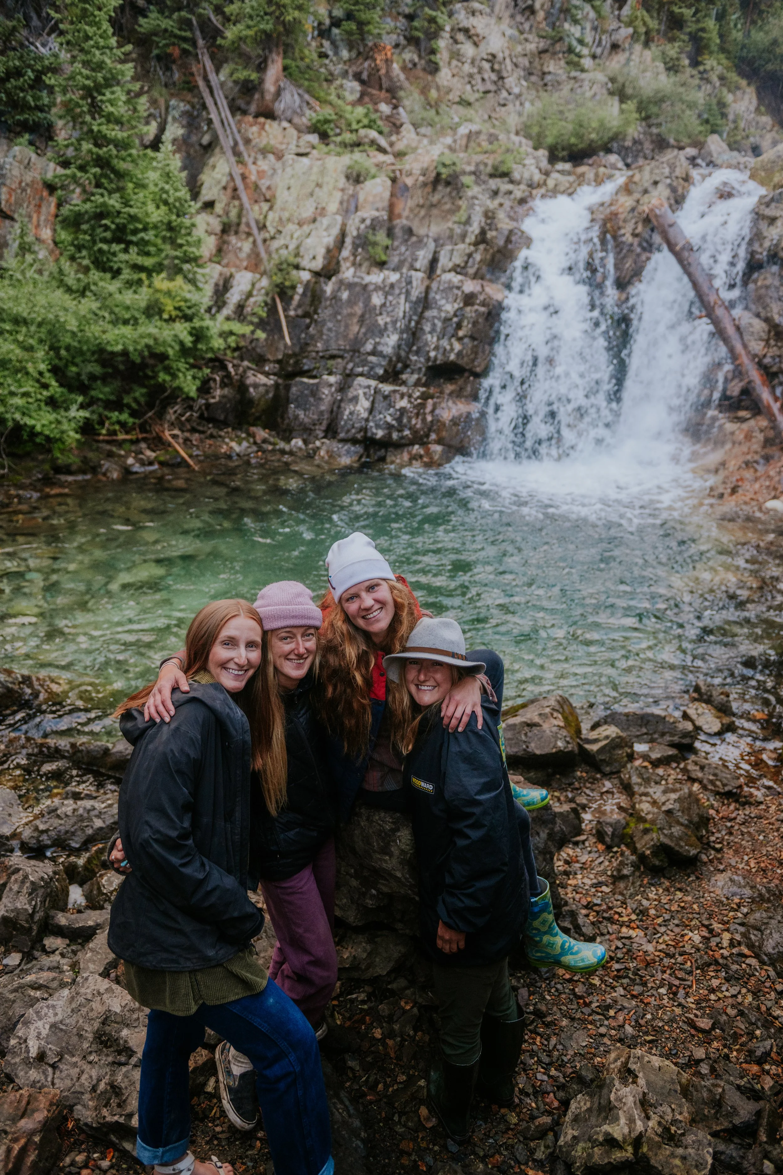 Group of five smiling women dressed in outdoor clothing, standing on rocky terrain near a waterfall in a forested area.