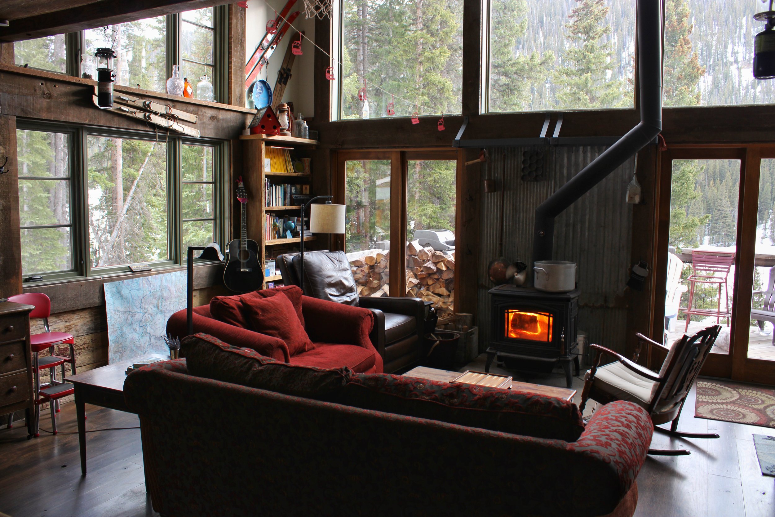 A cozy log cabin living room with large windows showing a forest outside. The room features a red patterned sofa, a black armchair, a wooden bookshelf filled with books, a guitar, a wood stove with a visible fire, a map on the wall, and various decorative items including bottles and skis.