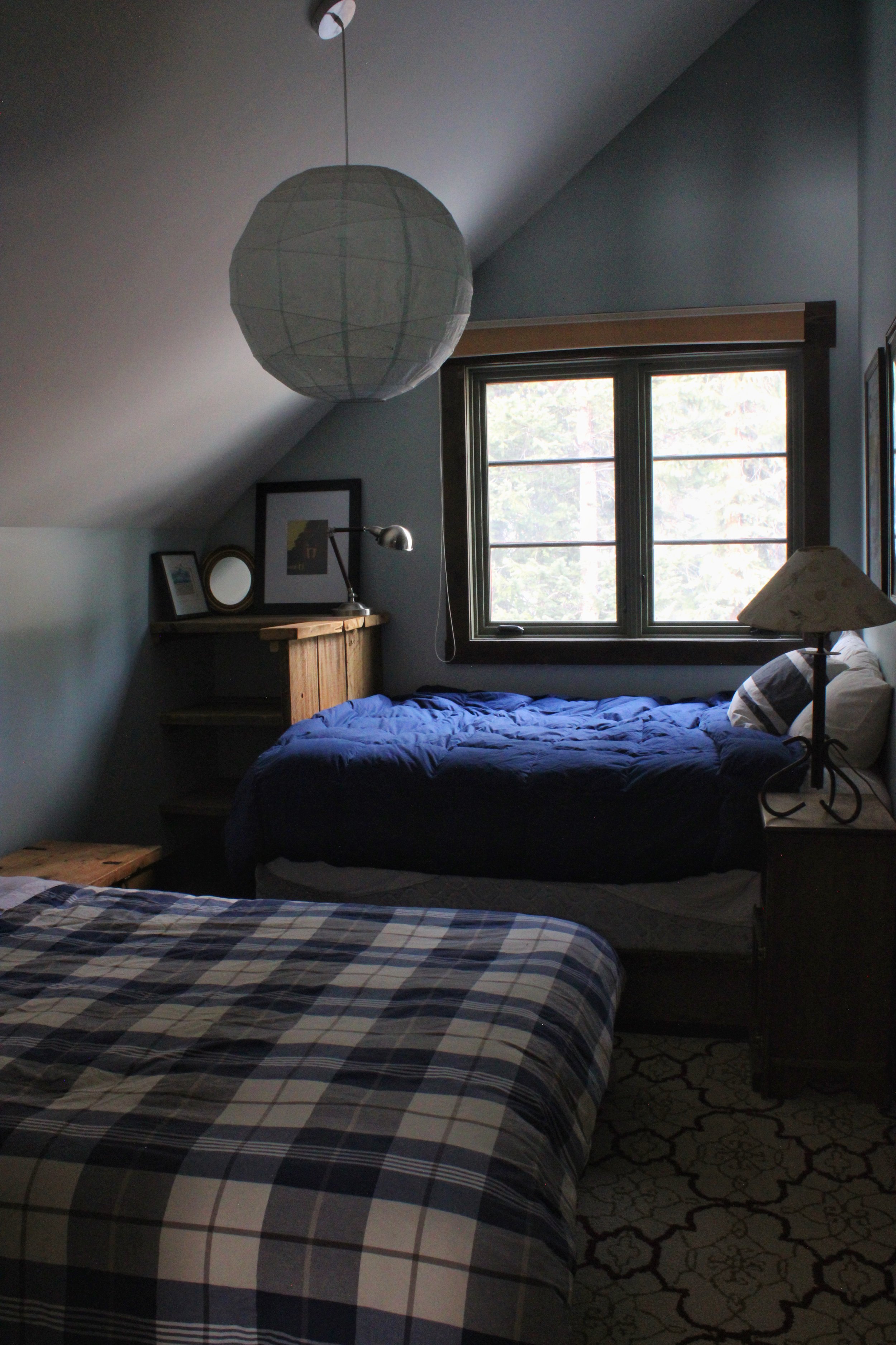 A bedroom with two beds, one in the foreground with a plaid pattern quilt, and another with a blue comforter. There is a window with view of trees, a wooden nightstand with a lamp, and a small shelf with pictures and a lamp. A paper lantern light fixture hangs from the ceiling.