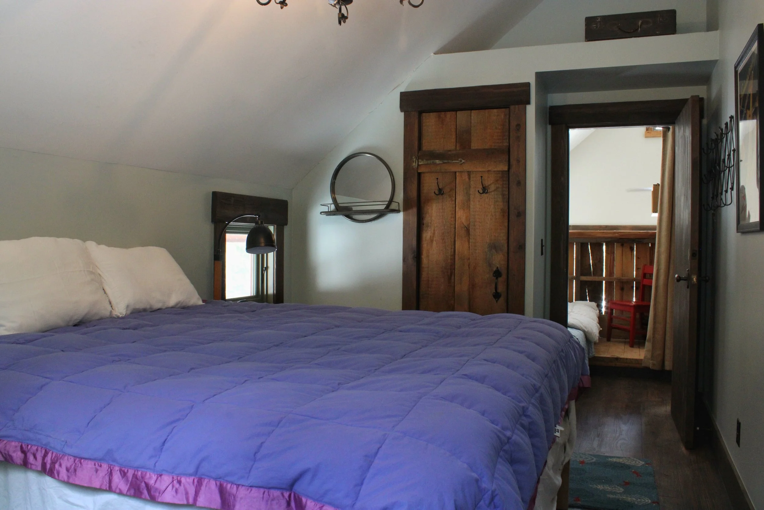 A cozy bedroom with a large bed covered in a purple quilt and white pillows, featuring a rustic wooden cabinet and a window with a black wall-mounted lamp next to it, with an open door leading to a balcony or outdoor area.