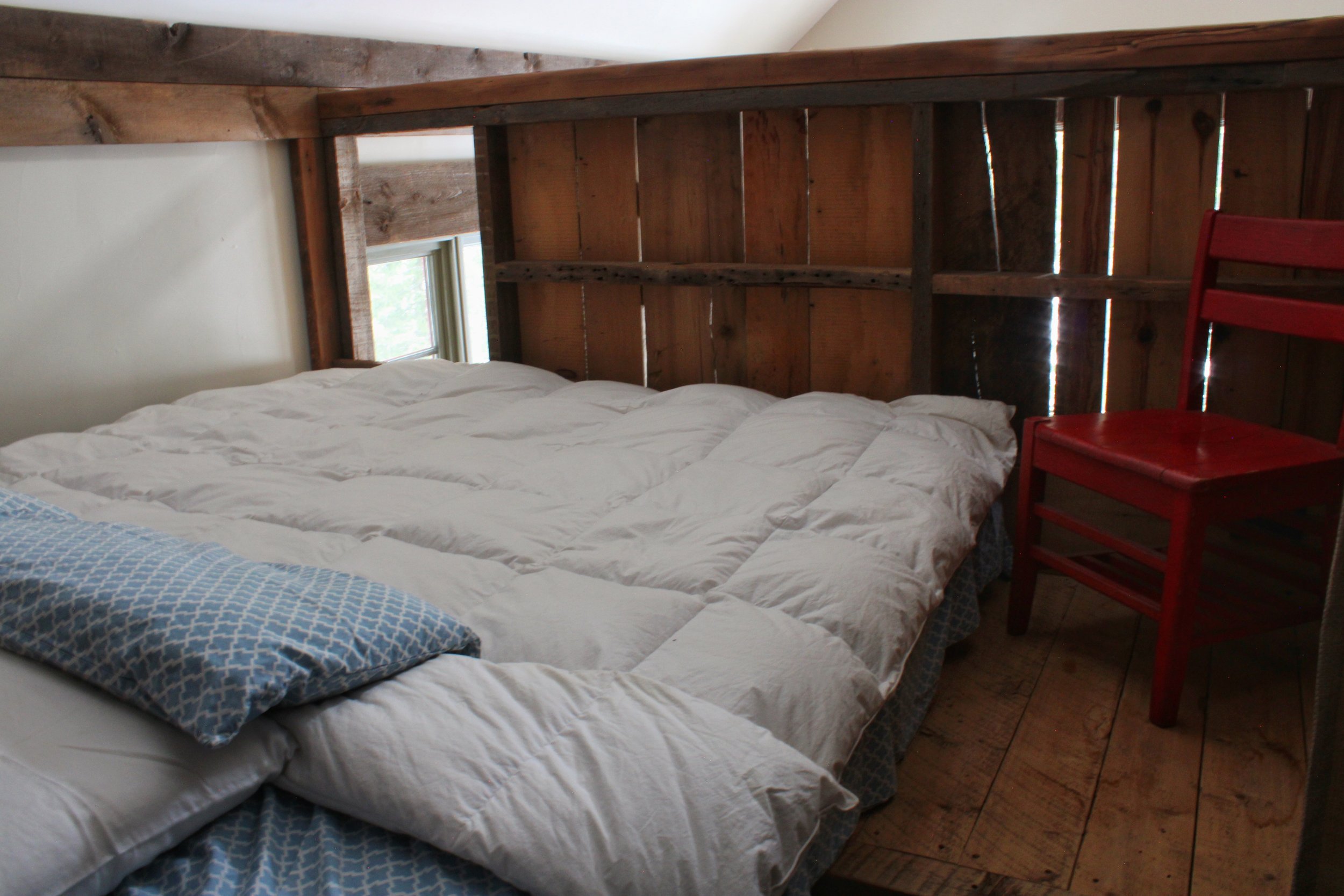 A bed with white comforter and blue patterned sheets in a rustic room with wooden walls and a window.