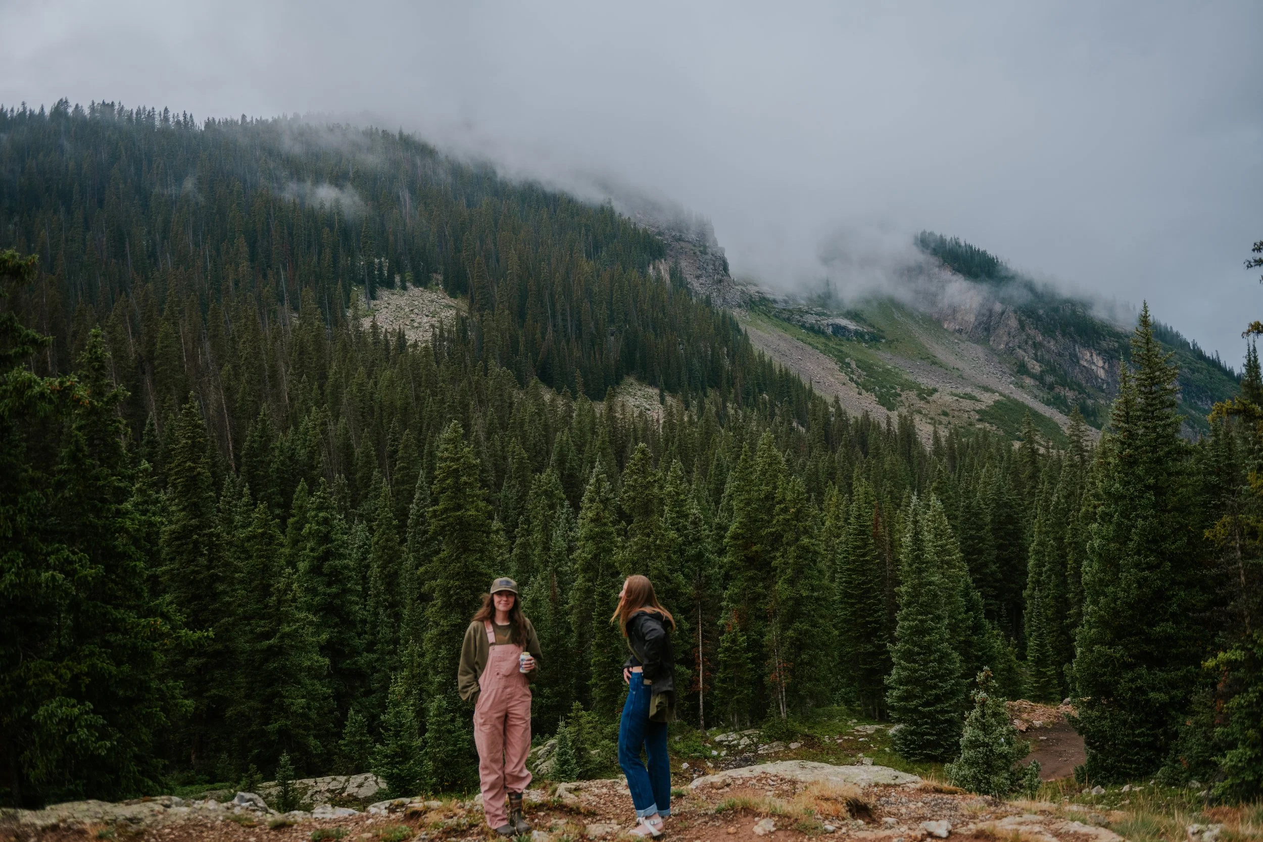 Three people standing on a rocky trail in a lush green forest with pine trees, mountain slopes, and low-hanging clouds in the background.