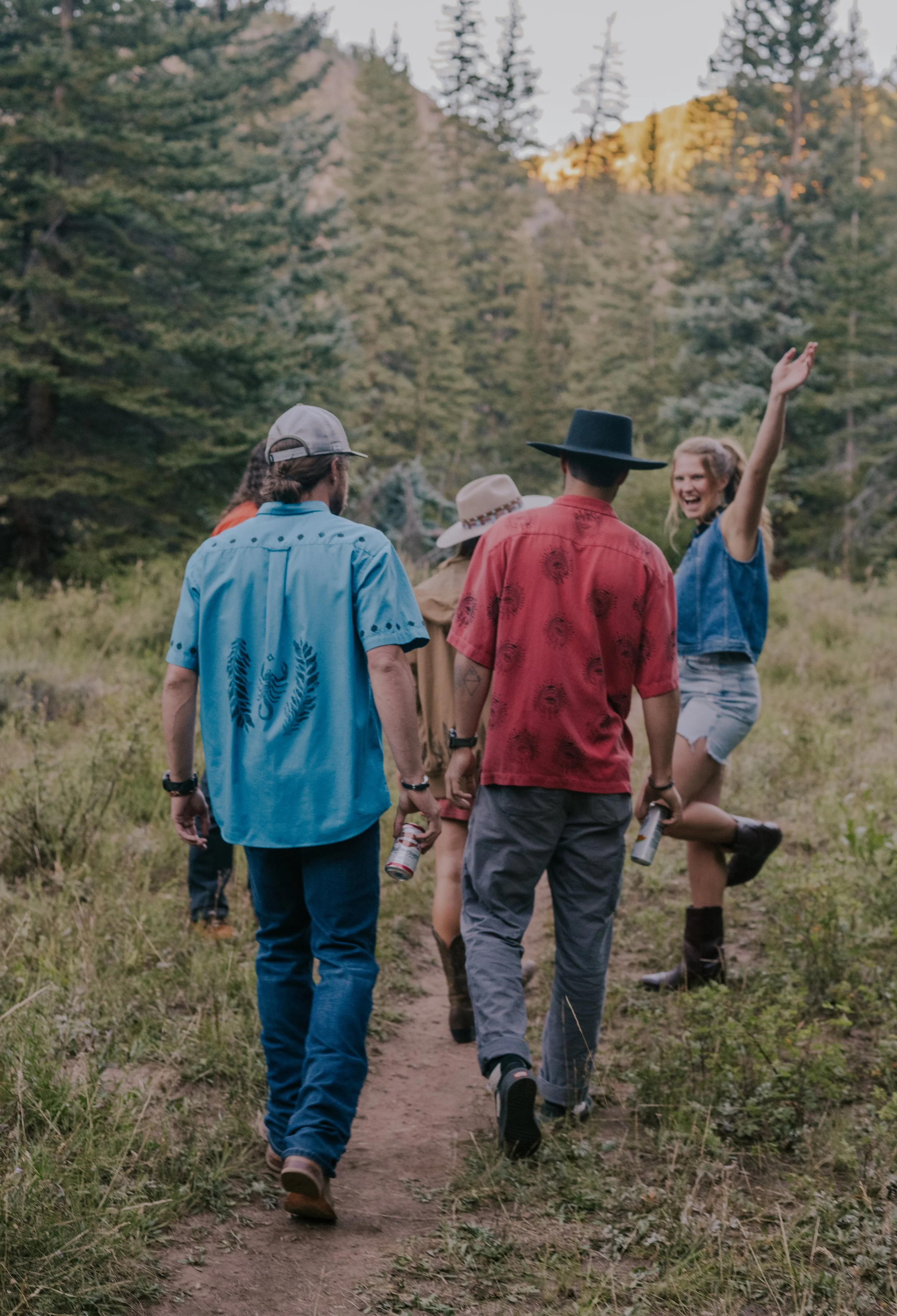 A group of five friends hiking on a trail in a forested area during daytime. They are walking away from the camera, with some carrying drinks and wearing casual summer clothes and hats. A young woman is smiling and waving at the camera.