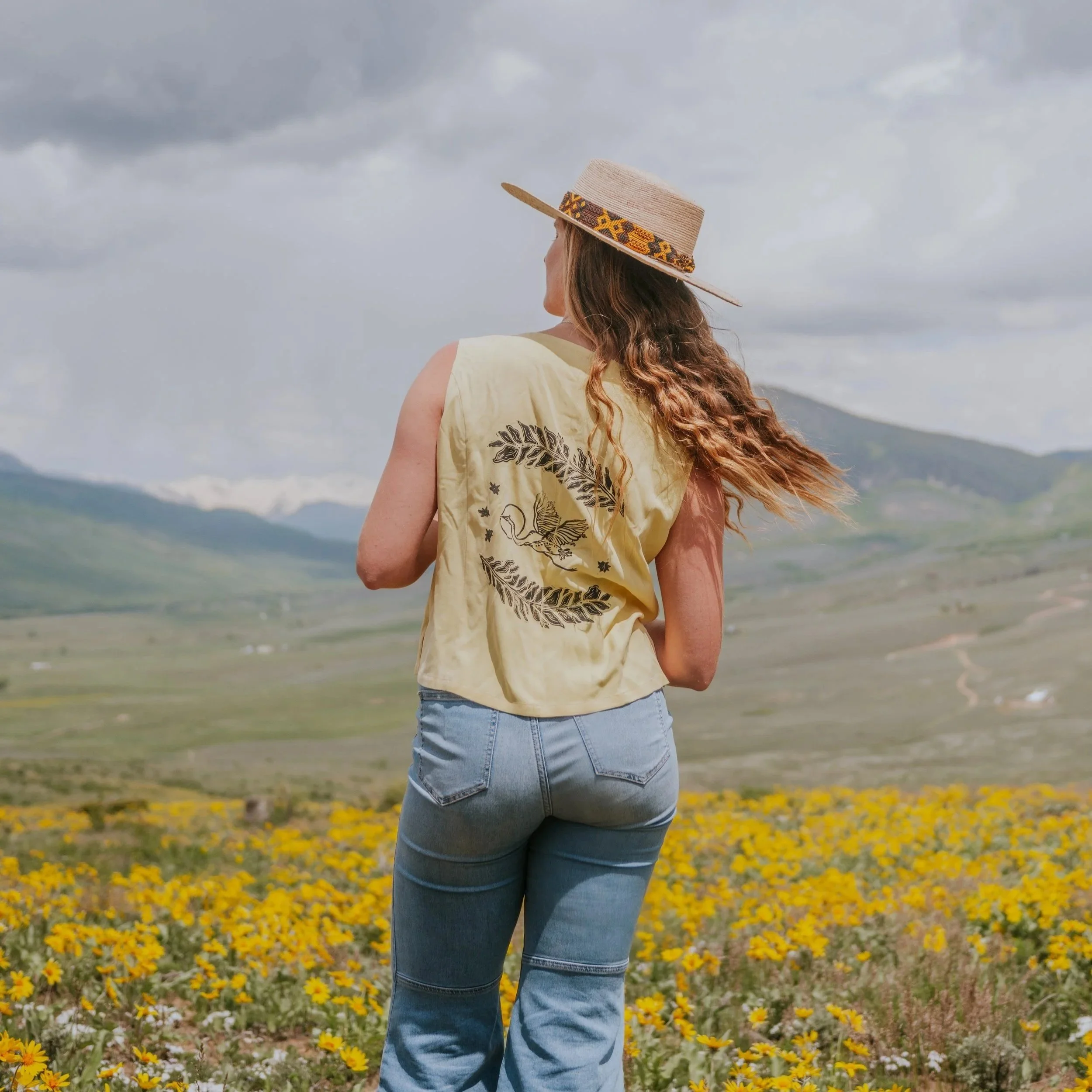 A woman with long wavy hair wearing a straw hat with decorative band, a beige sleeveless top, and blue jeans, standing in a field of yellow flowers with mountains and cloudy sky in the background.