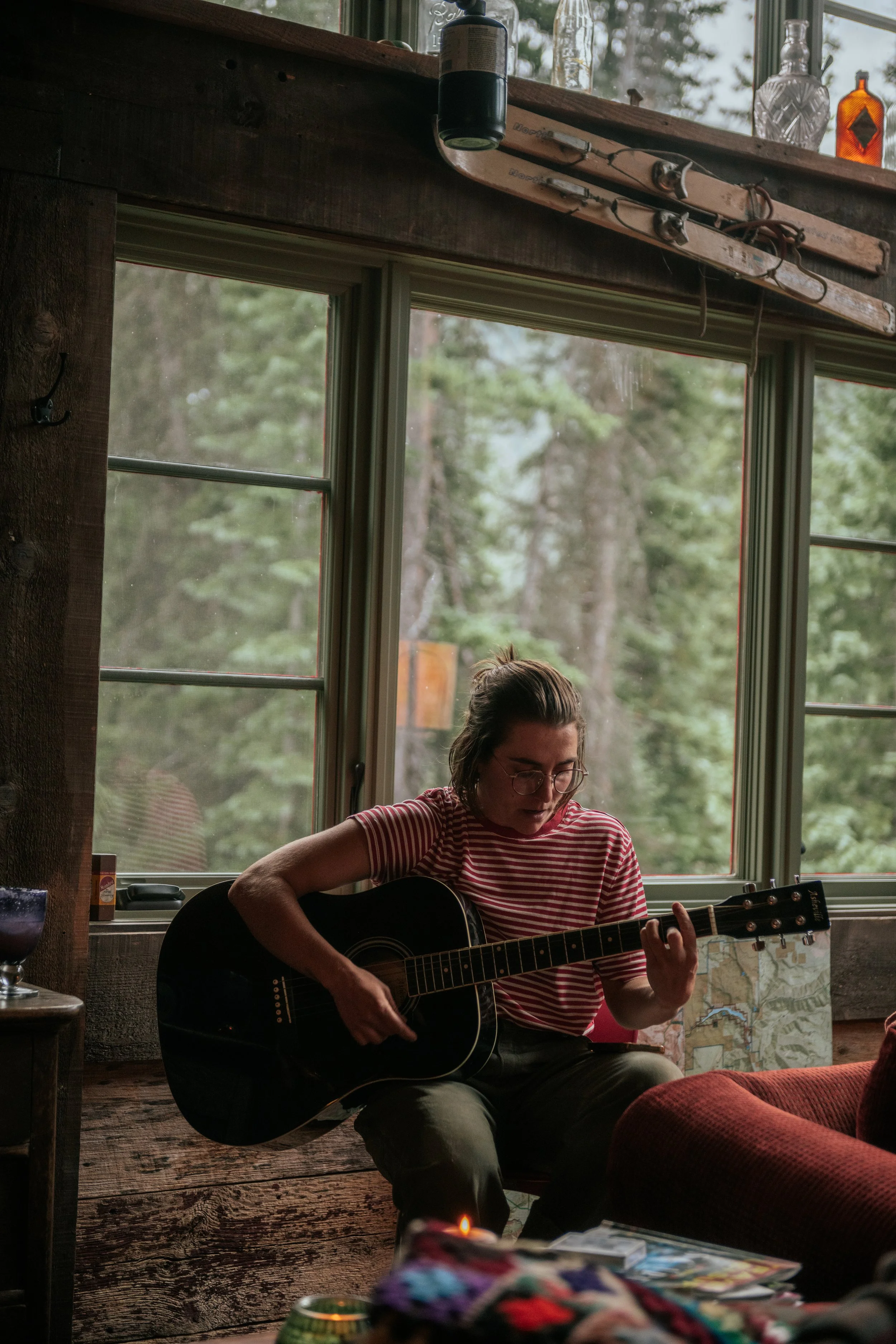 A young woman with glasses and a striped shirt is sitting by a window, playing an acoustic guitar. The room has a rustic wooden interior with a large window showing trees outside. There are bottles and decorative items on a shelf above the window.