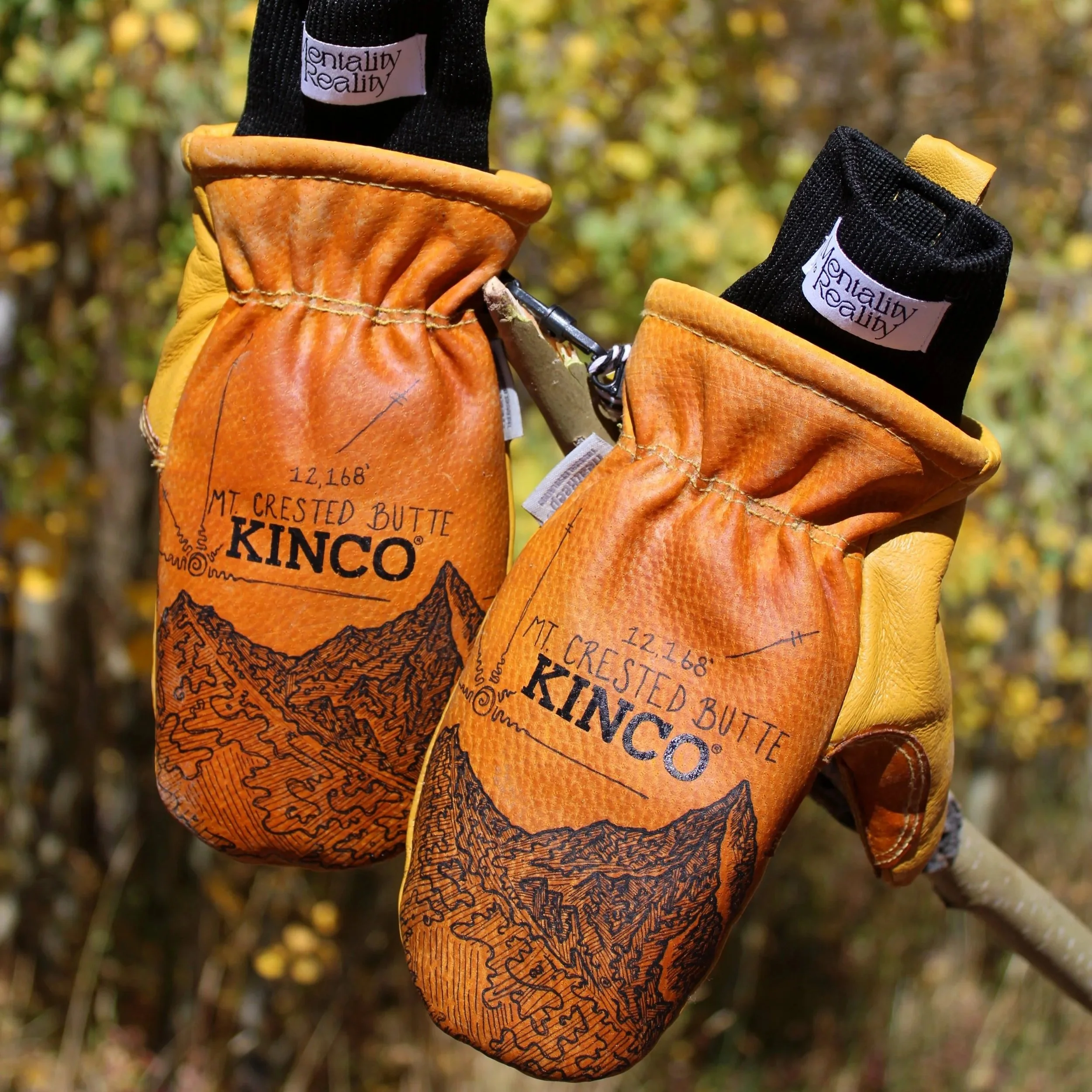 Close-up of yellow leather gloves with black fabric cuffs, featuring the brand label 'Mentality Reality' and a scenic mountain map design on the leather, outdoors with blurred autumn foliage in the background.