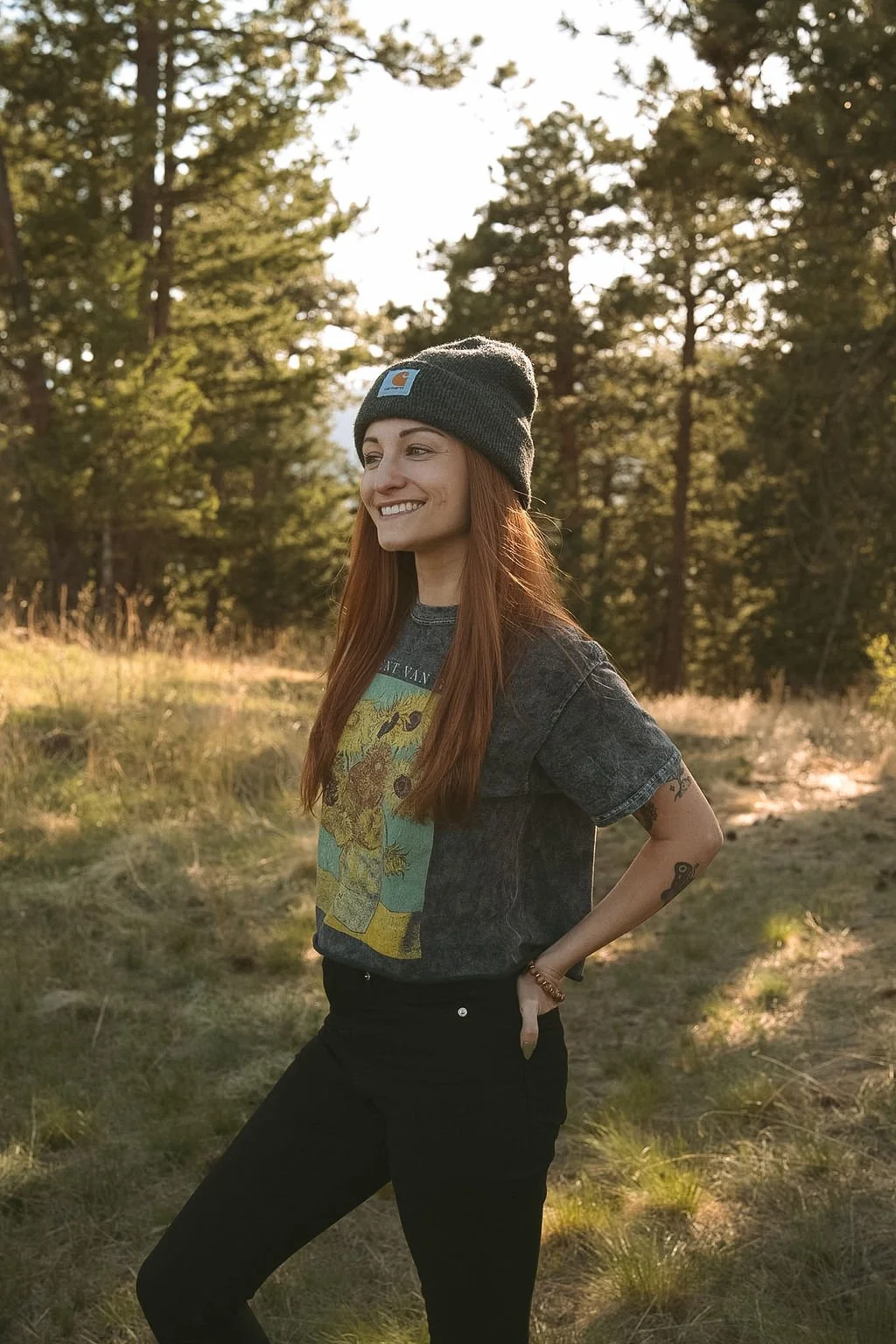 A woman outdoors in a forest setting, smiling and looking to the side. She is wearing a dark beanie, a graphic T-shirt, and black pants.