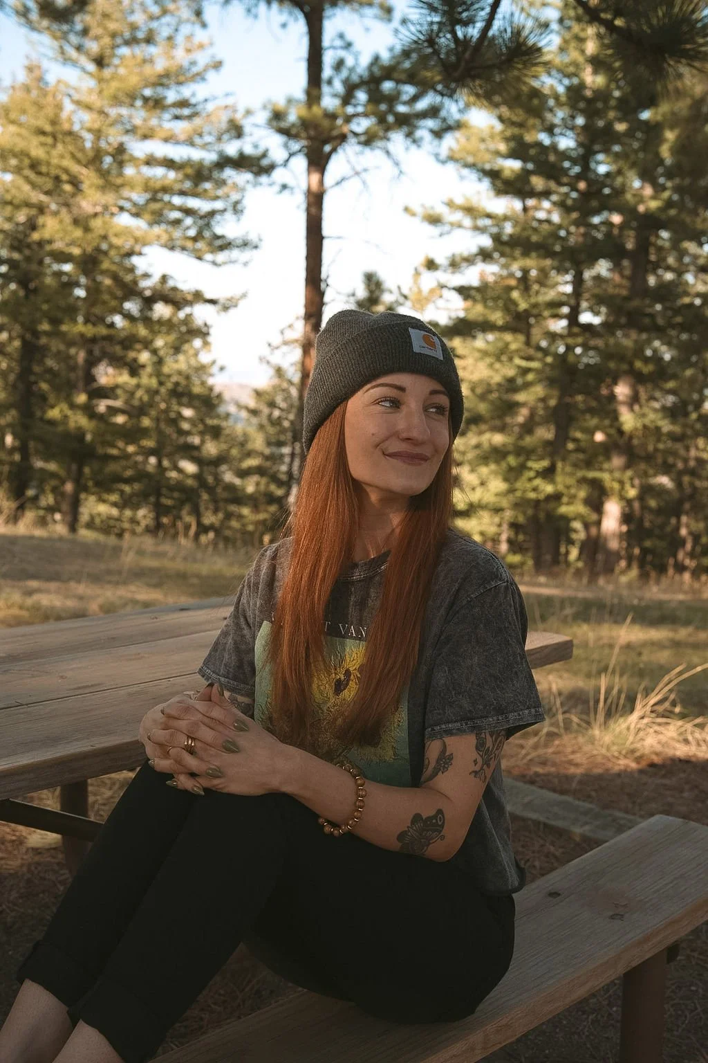 A woman with red hair, wearing a gray beanie and a black graphic t-shirt, sitting on a wooden bench outdoors in a forested area during the day.