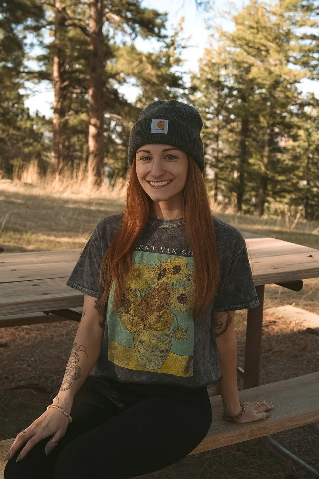 A young woman with long red hair, wearing a gray beanie, a graphic T-shirt, and black pants, sitting on a wooden bench outdoors in a forested area with trees in the background, smiling at the camera.
