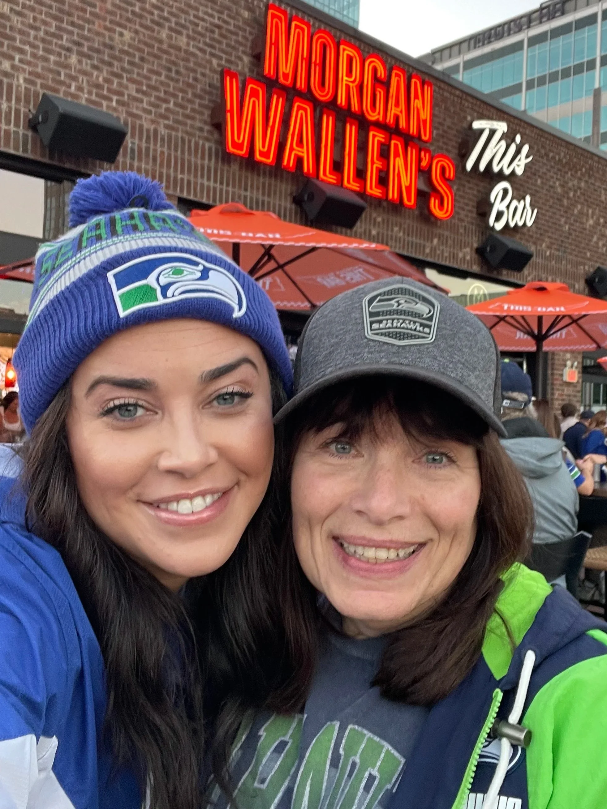 Two women at a bar outside Morgan Wallen's bar, smiling and wearing Seattle Seahawks hats and jackets, with outdoor umbrellas and a neon sign for Morgan Wallen's bar in the background.