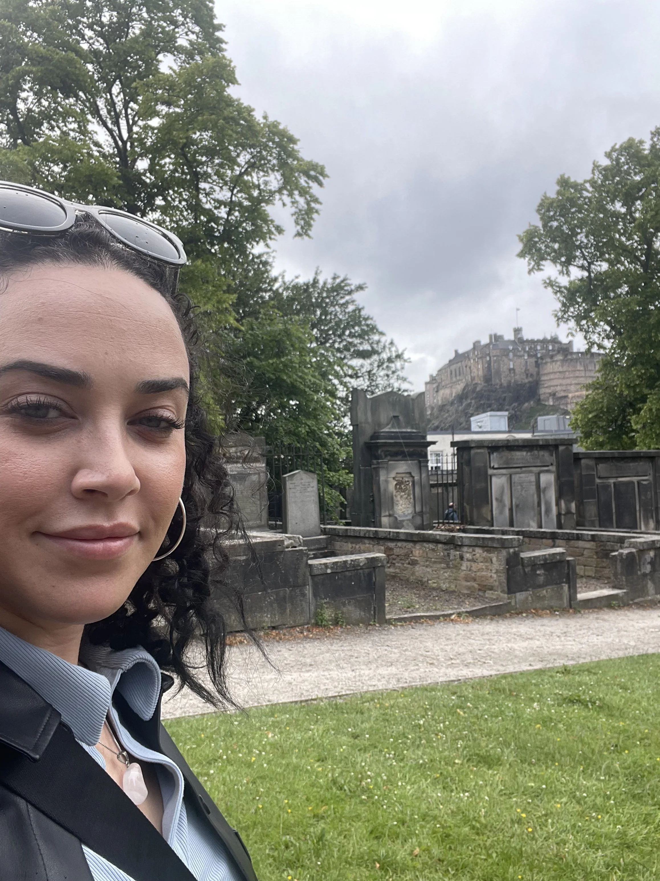 A woman with dark curly hair and sunglasses on her head, standing in front of a historical site with a castle on a hill, trees, stone walls, and a graveyard or memorial with stone tombs. Overcast sky.