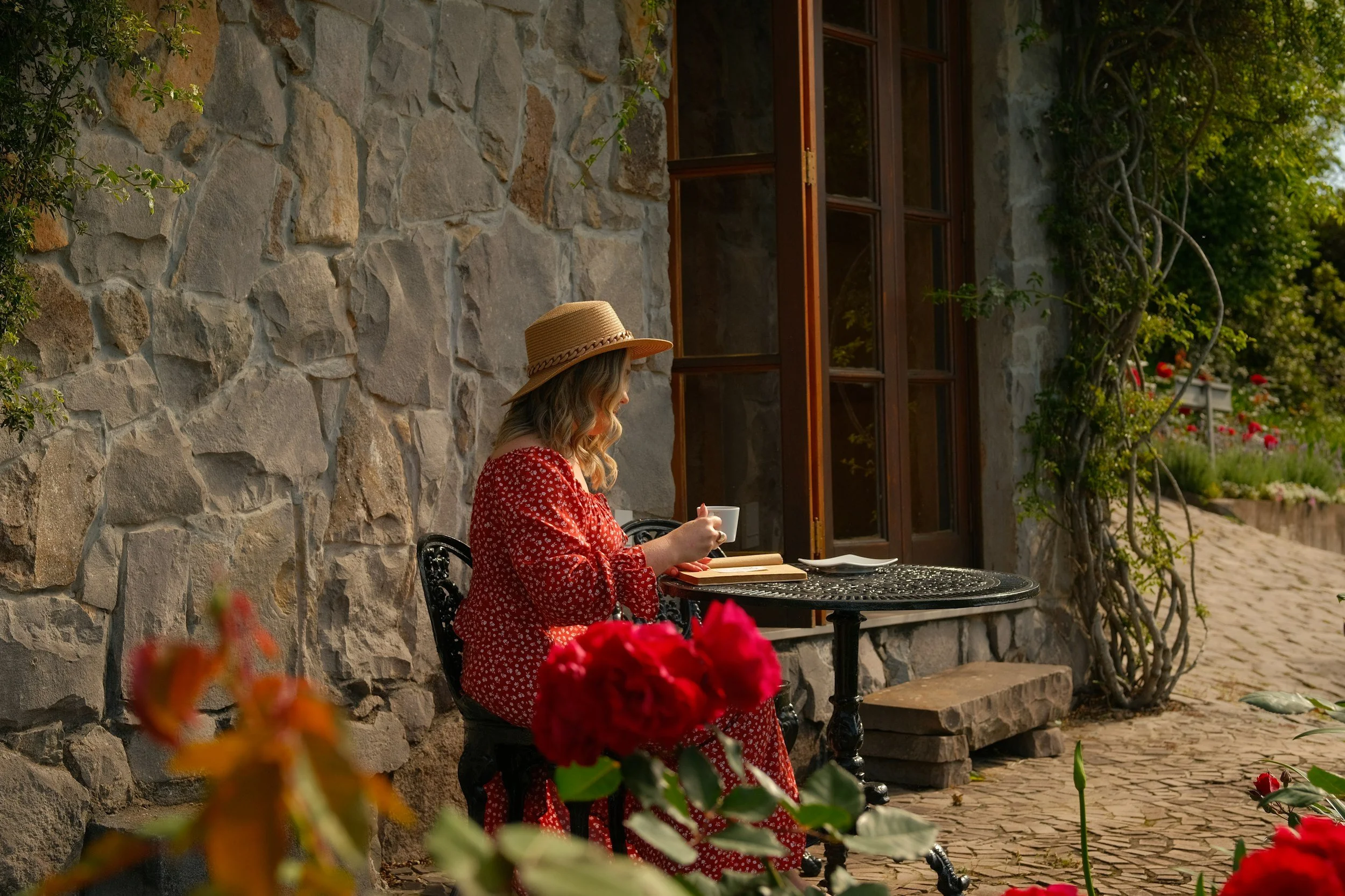 A Scandinavian wellness scene featuring a midlife woman in a red patterned dress, sitting outdoors with a journal and coffee beside a stone wall and blooming roses. The image reflects Nordic ritual writing, quiet reflection, and the grounded clarity