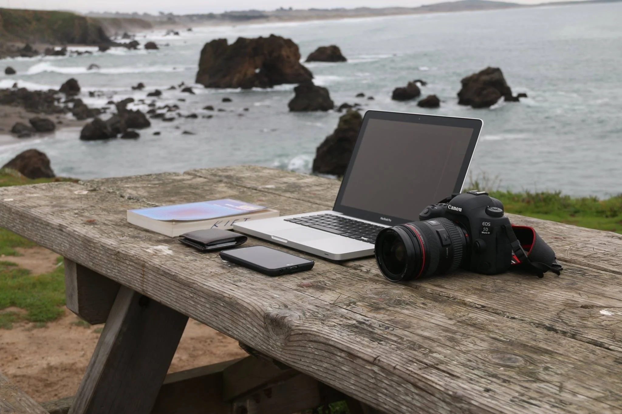 A Nordic coastal workspace with a laptop, camera, and journal set on a wooden table overlooking rugged rocks and ocean waves. The scene reflects Scandinavian creative work, midlife storytelling, and the grounded clarity behind Sissel Bridges’ media a