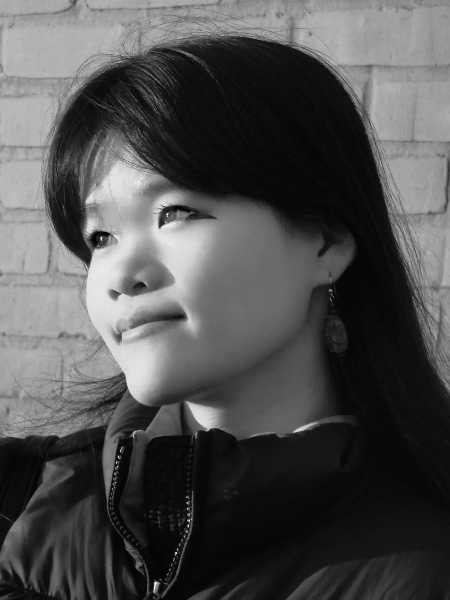 Close-up black and white photo of a young person with dark hair, looking to the side, wearing a jacket with a visible zipper and studded collar, against a brick wall background.