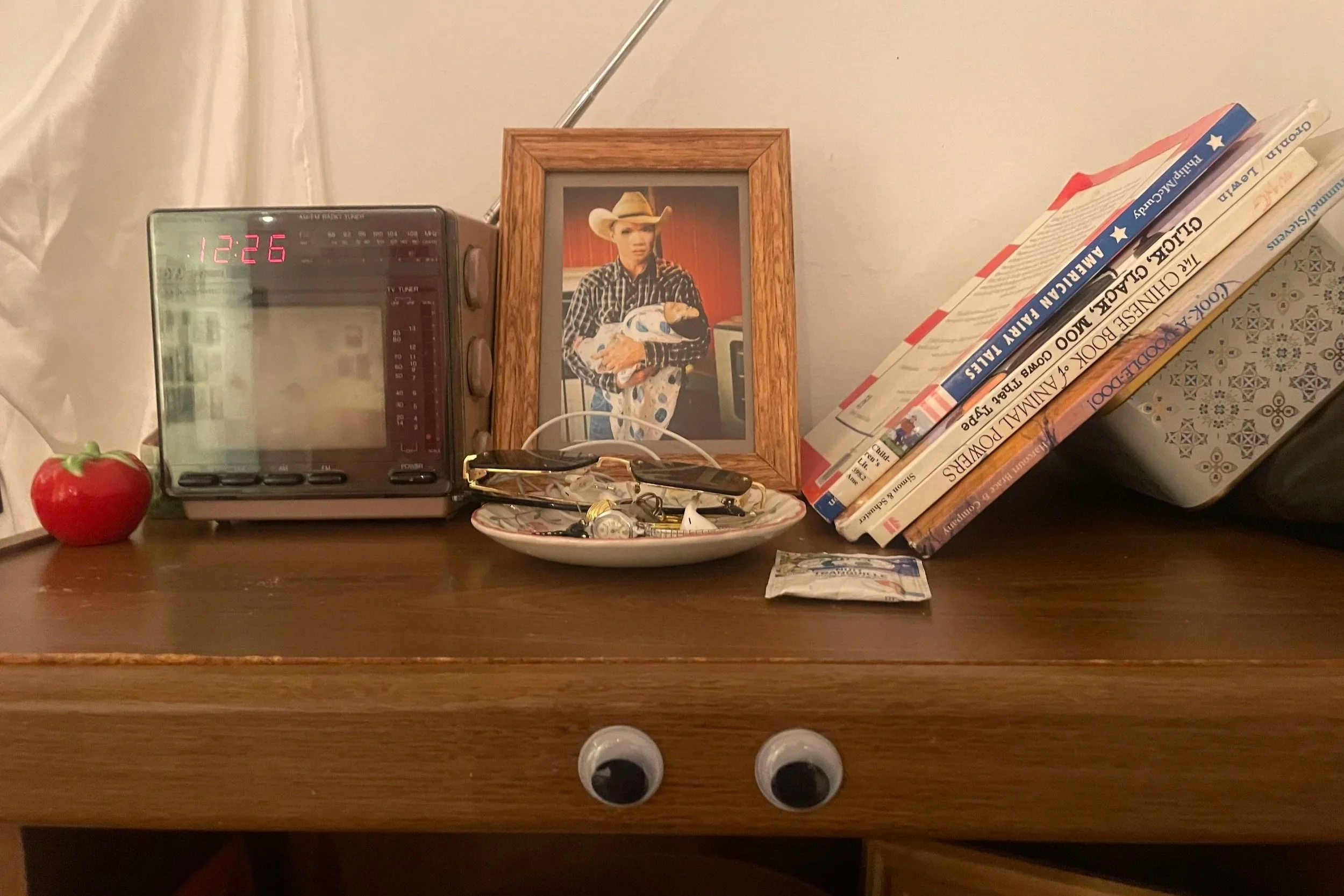 A wooden table with a red apple, a small clock radio displaying 12:26, a framed photo of a woman wearing a cowboy hat and plaid shirt holding a patterned cloth, a cup of glasses, a small bowl with miscellaneous items, several stacked books including titles related to country music, a small paper note, an electrical cord, and a pair of googly eyes.