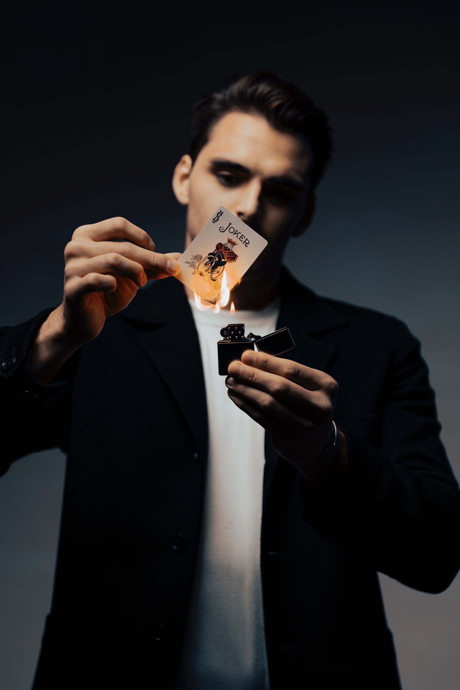 A man in a black blazer and white shirt is holding a burning playing card with a Joker design and lighting a lighter in a dark setting.