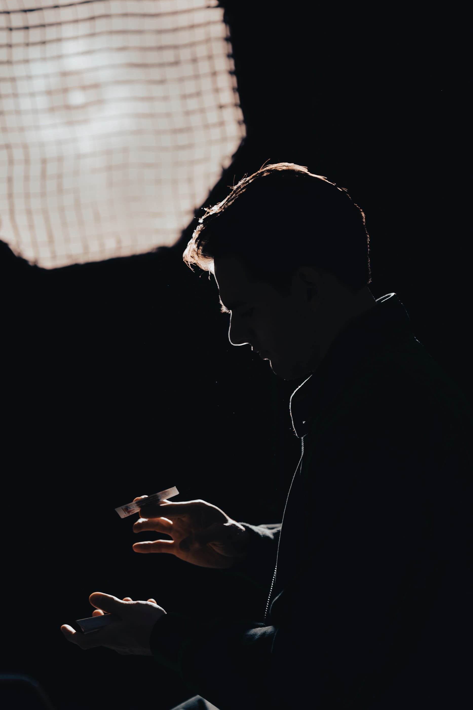 Silhouette of a person looking at a phone, backlit by a light-colored window or screen, with visible earphones and holding a credit card in hand, set against a dark background.