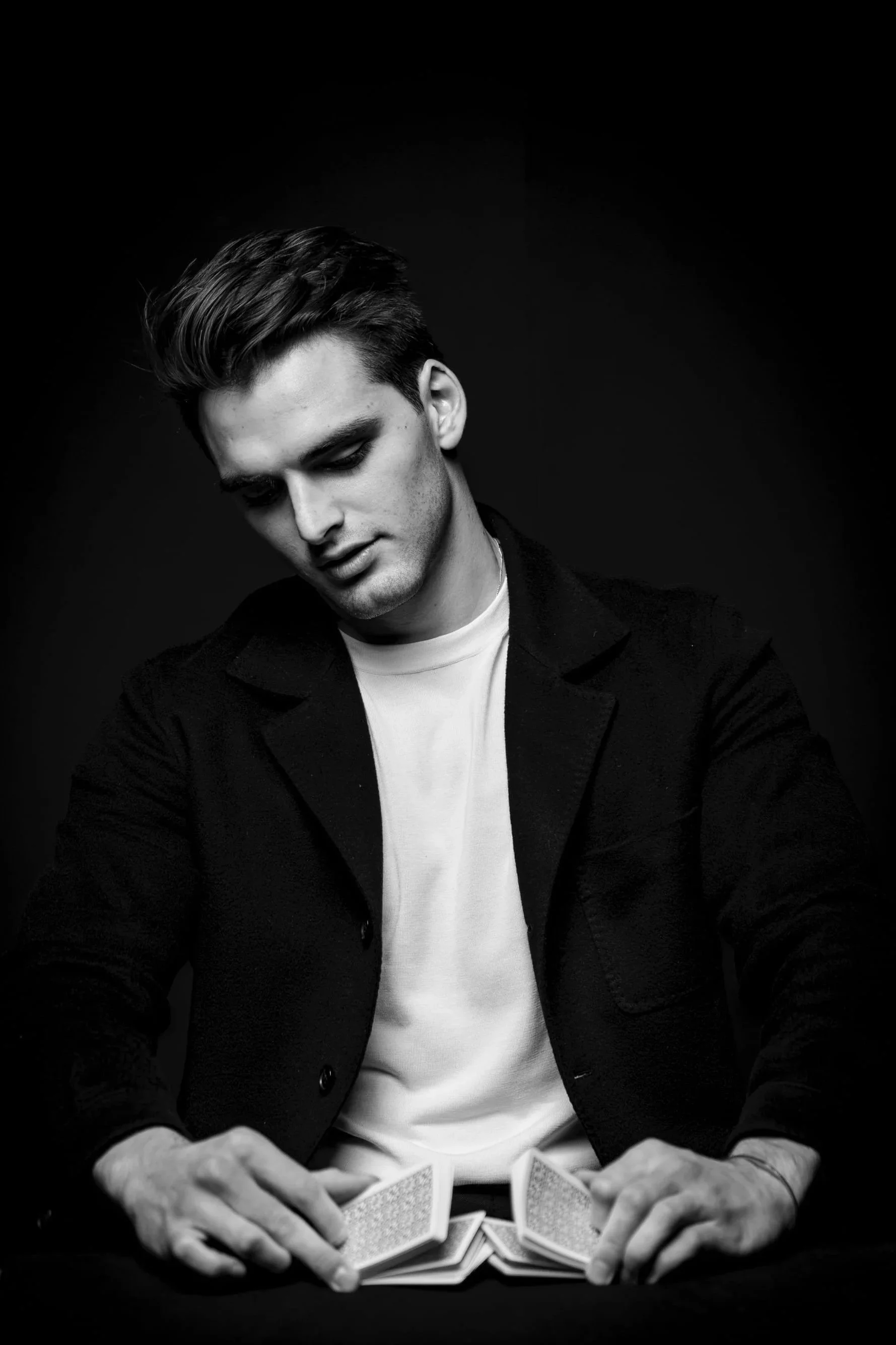 Black and white photo of a young man with styled hair, seated at a table, shuffling a deck of playing cards. He is wearing a white t-shirt and a dark blazer, with a neutral expression, looking down at the cards.