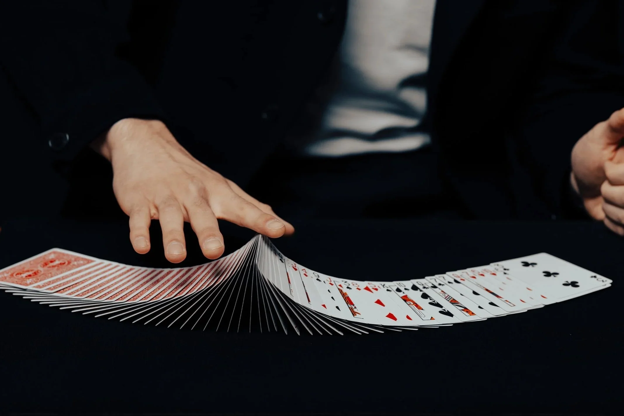 A person in a dark jacket and white shirt is flipping playing cards across a black surface, with a spread of cards laid out on the table.